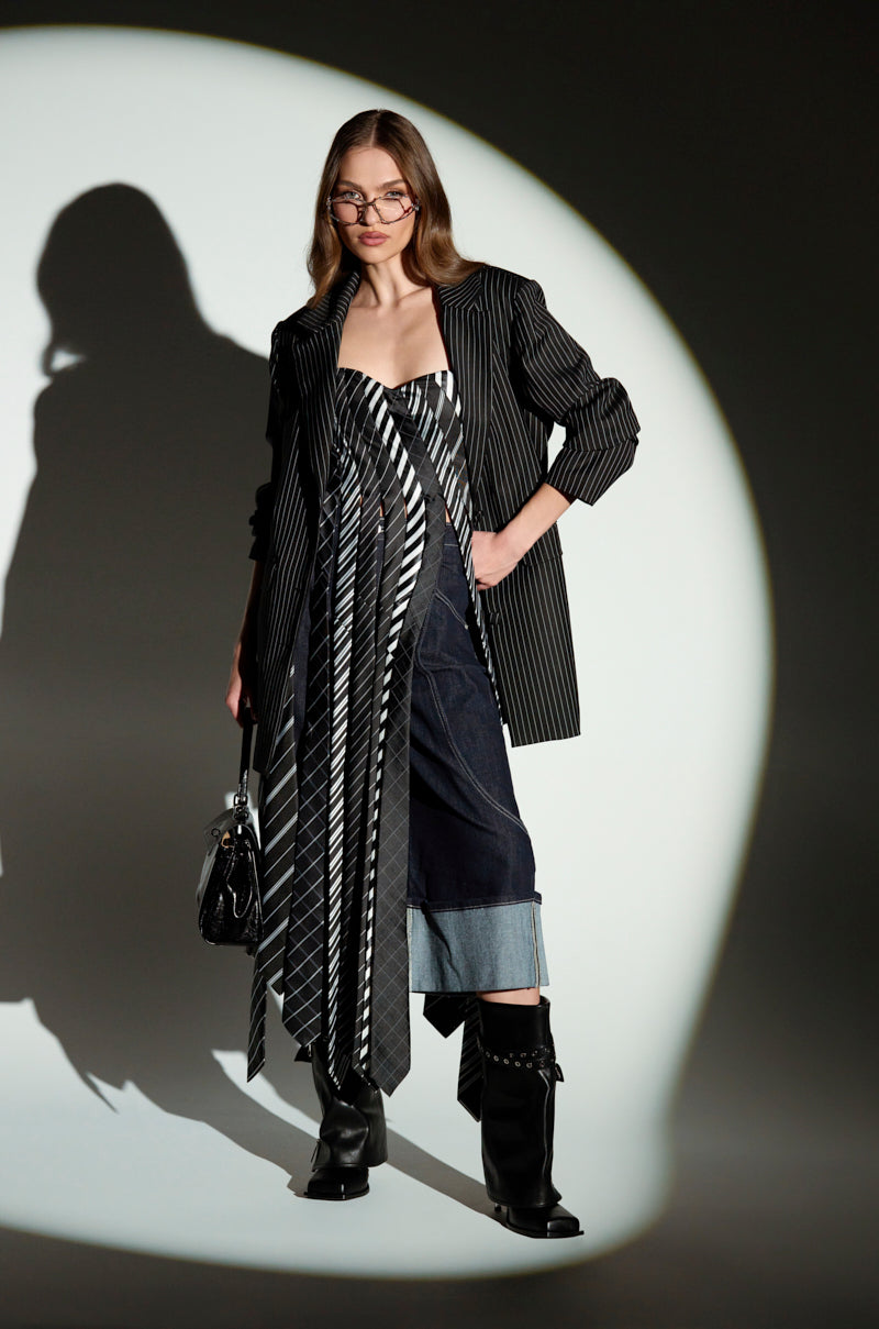 A woman confidently dons a pinstripe blazer over the ALL WORK AND NO PLAY TIE DETAIL CORSET TOP. Her long, wavy hair and glasses complement her look, finished with cuffed jeans, black boots, and a handbag, all under a spotlight.
