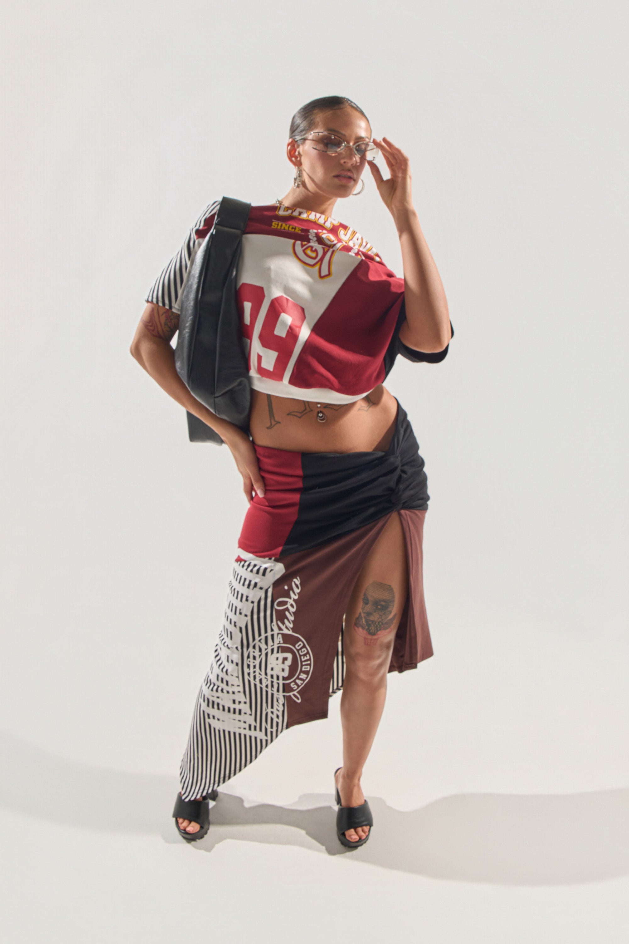 A woman poses against a white background in the SCHOOL SPIRIT REWORKED MIDI SKIRT, styled with bold colors and prints, black heeled sandals, glasses, and a jacket draped over one shoulder for a sporty, patchwork-inspired look. 3