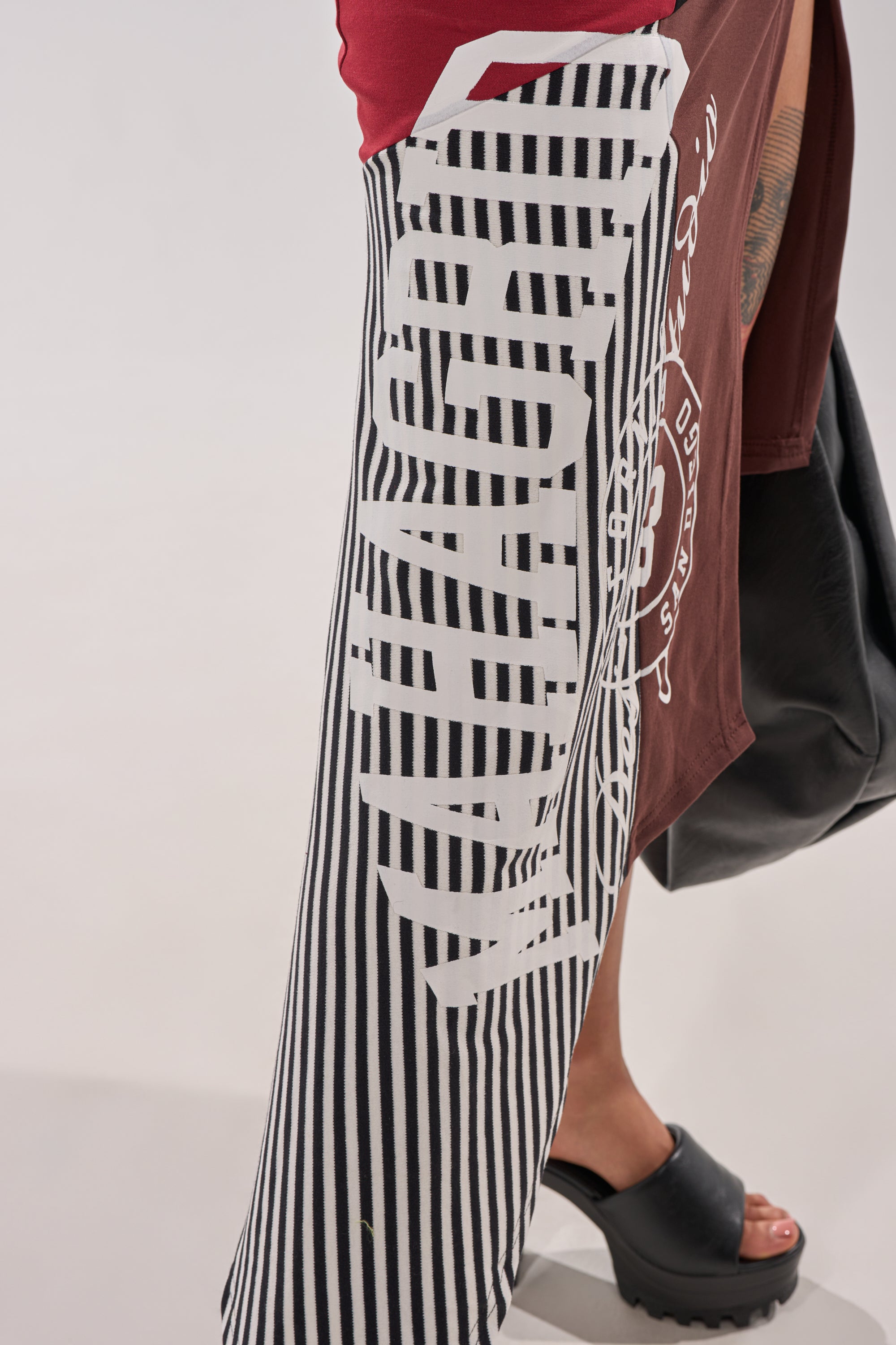 Someone is wearing the SCHOOL SPIRIT REWORKED MIDI SKIRT, which has a high-rise wrap front and sporty stripe paneling, paired with a maroon white-print top and black platform sandals. Only part of their lower body is shown against a plain background. 10