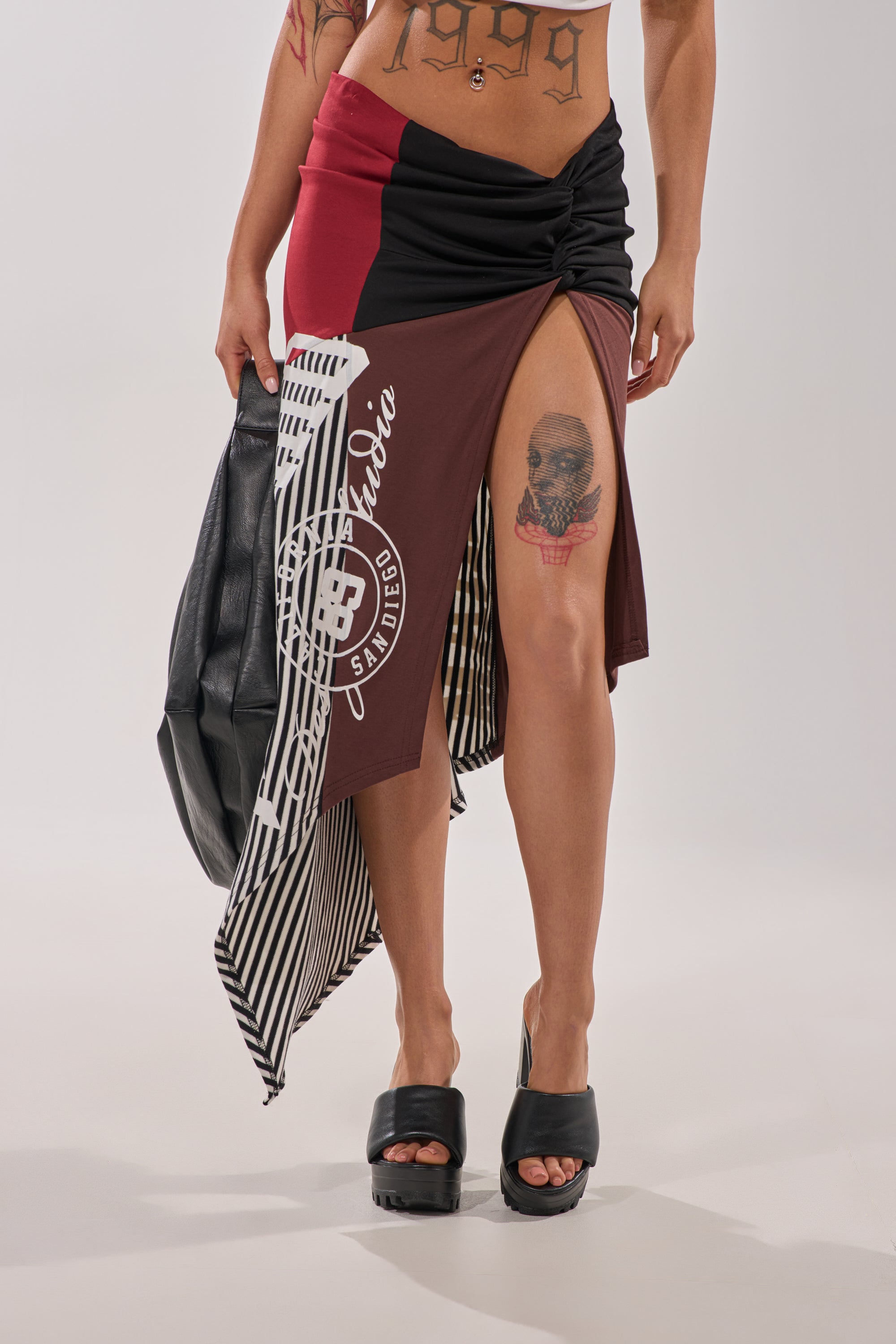Wearing the SCHOOL SPIRIT REWORKED MIDI SKIRT with bold prints and sporty stripes, paired with black platform sandals and a black bag. Tattoos are visible on their thigh and torso. 0