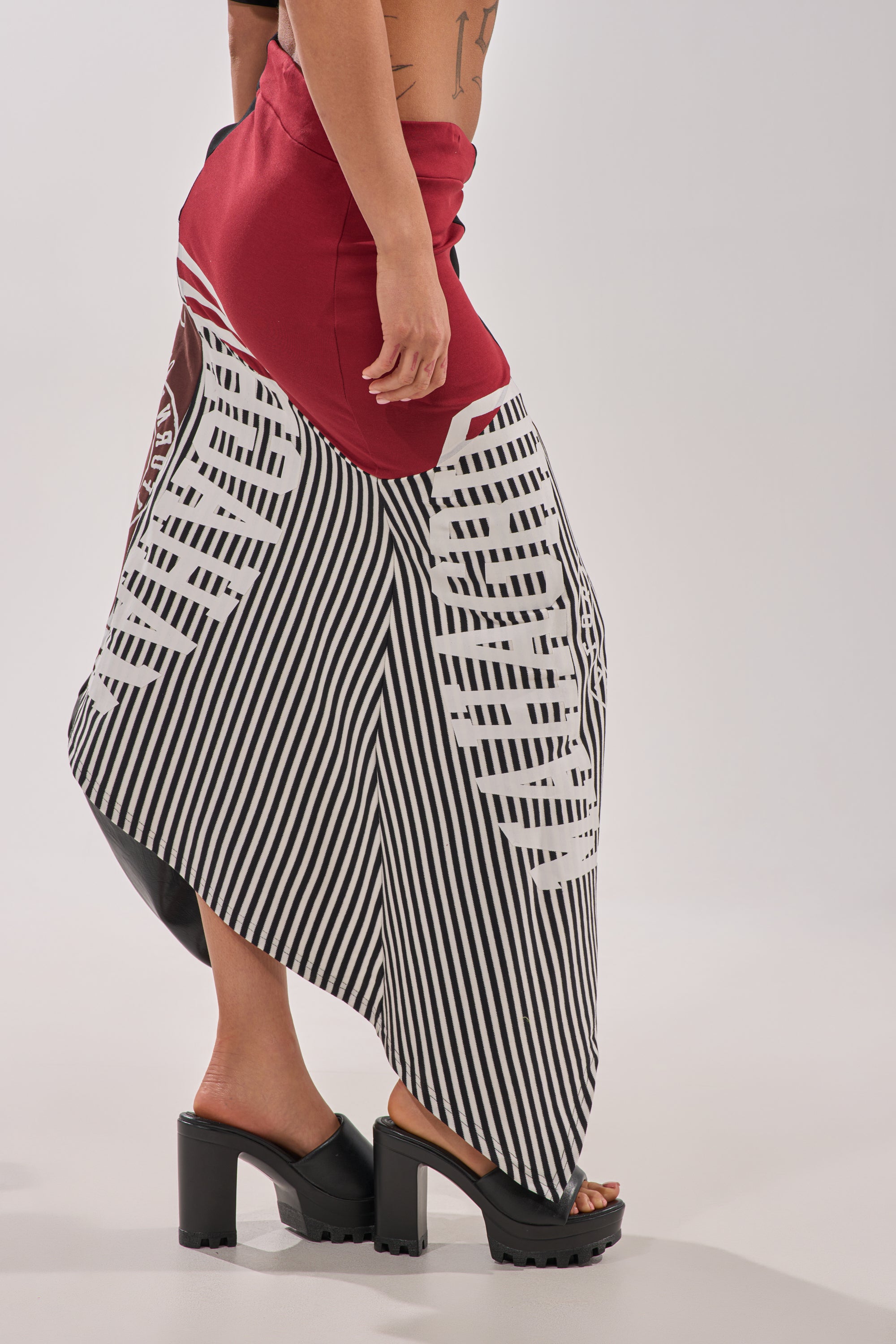 Someone wears the SCHOOL SPIRIT REWORKED MIDI SKIRT—a red midi skirt with a black-and-white striped geometric overlay—paired with chunky black platform heels, standing in front of a plain background. 7