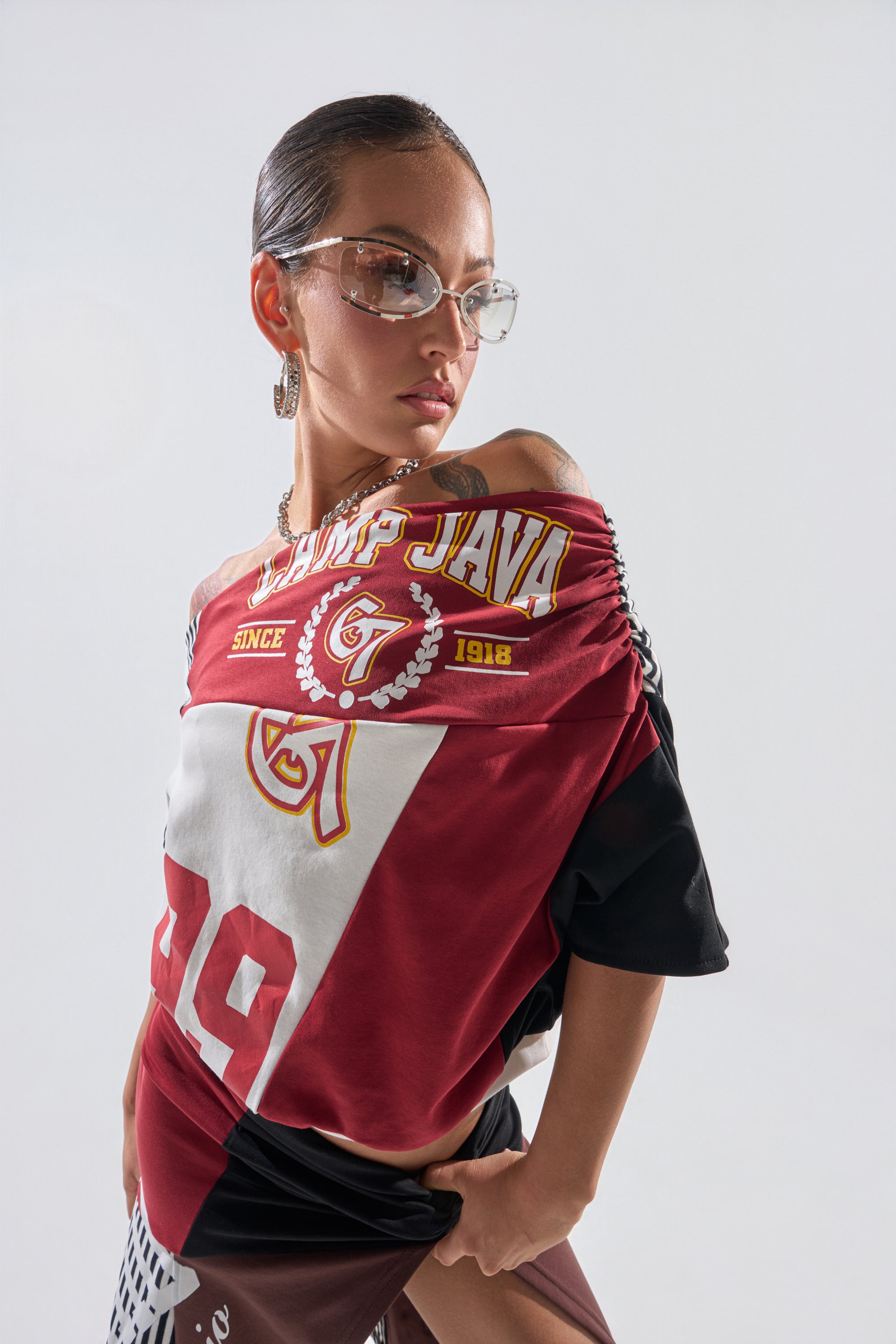 A woman wearing clear glasses and the SCHOOL SPIRIT REWORKED TEE—a red varsity-style off-the-shoulder graphic top—poses confidently with her hand on her hip against a plain white background. 0