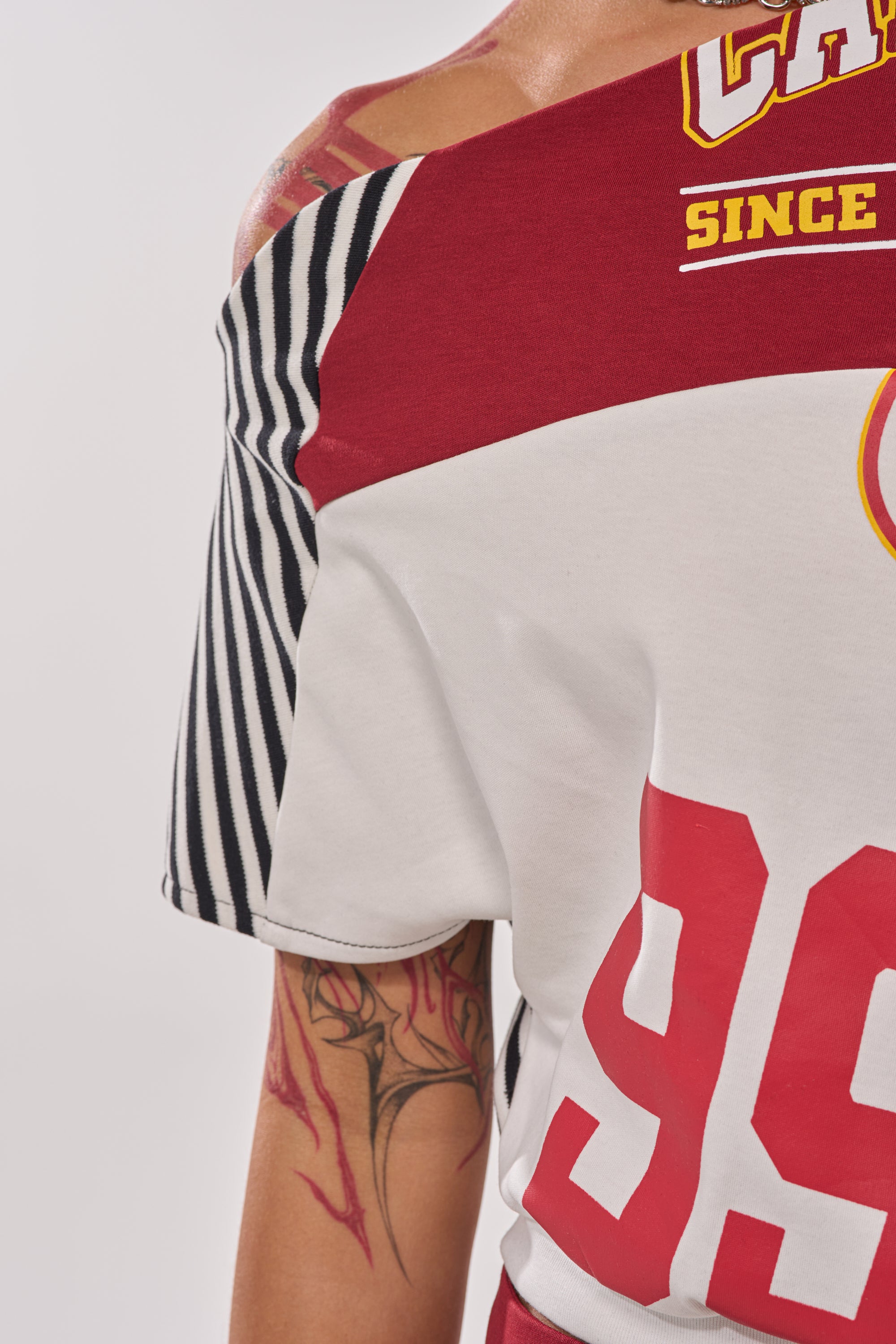 Wearing the SCHOOL SPIRIT REWORKED TEE—an off-the-shoulder red, white, and black striped graphic top with bold text—a person shows off their colorful flame tattoo for standout streetwear style. 10