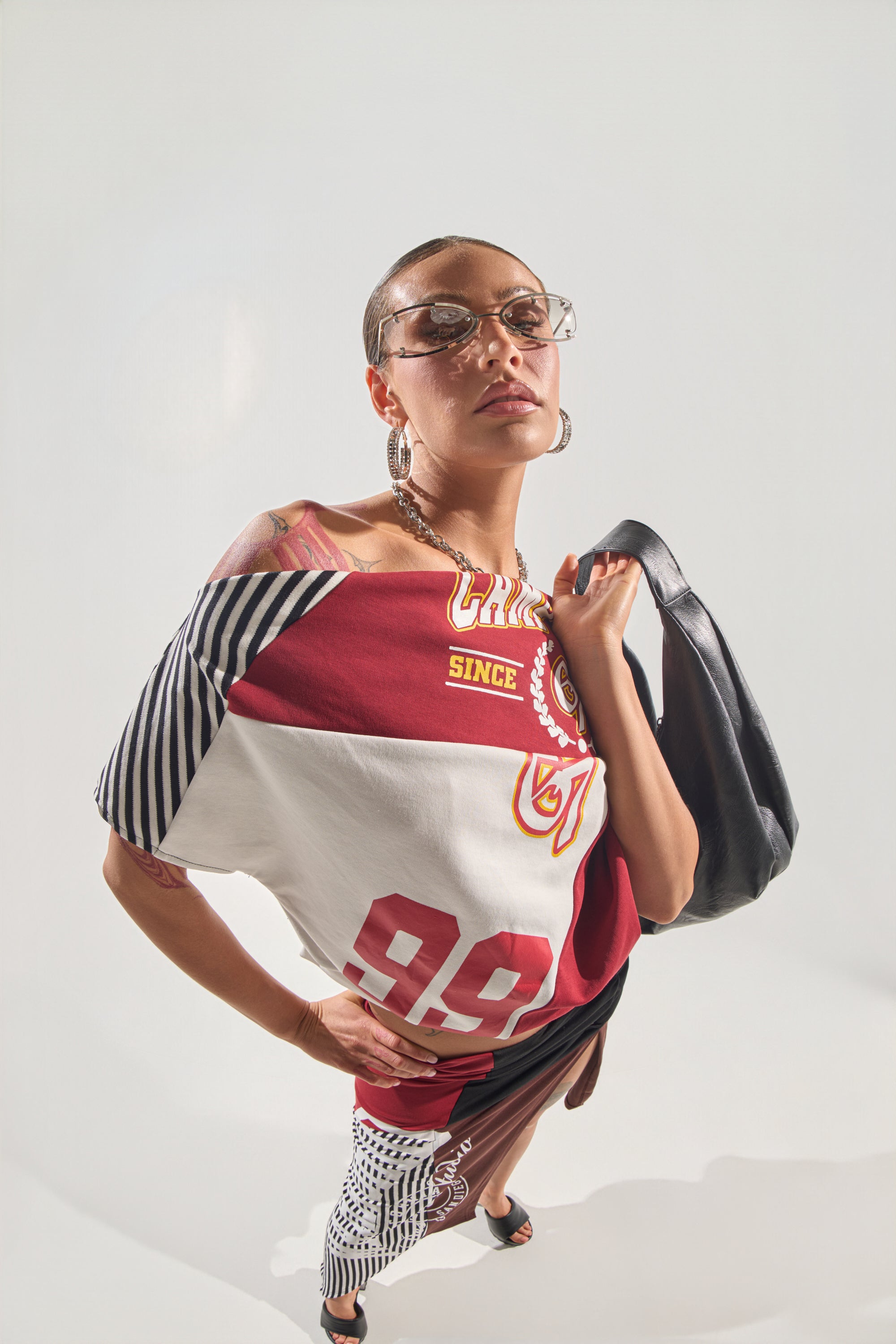A confident person wears the SCHOOL SPIRIT REWORKED TEE—a mixed-pattern varsity-style top with numbers and stripes—paired with glasses, hoop earrings, and a black bag over one shoulder, looking down at the camera against a white background. 1