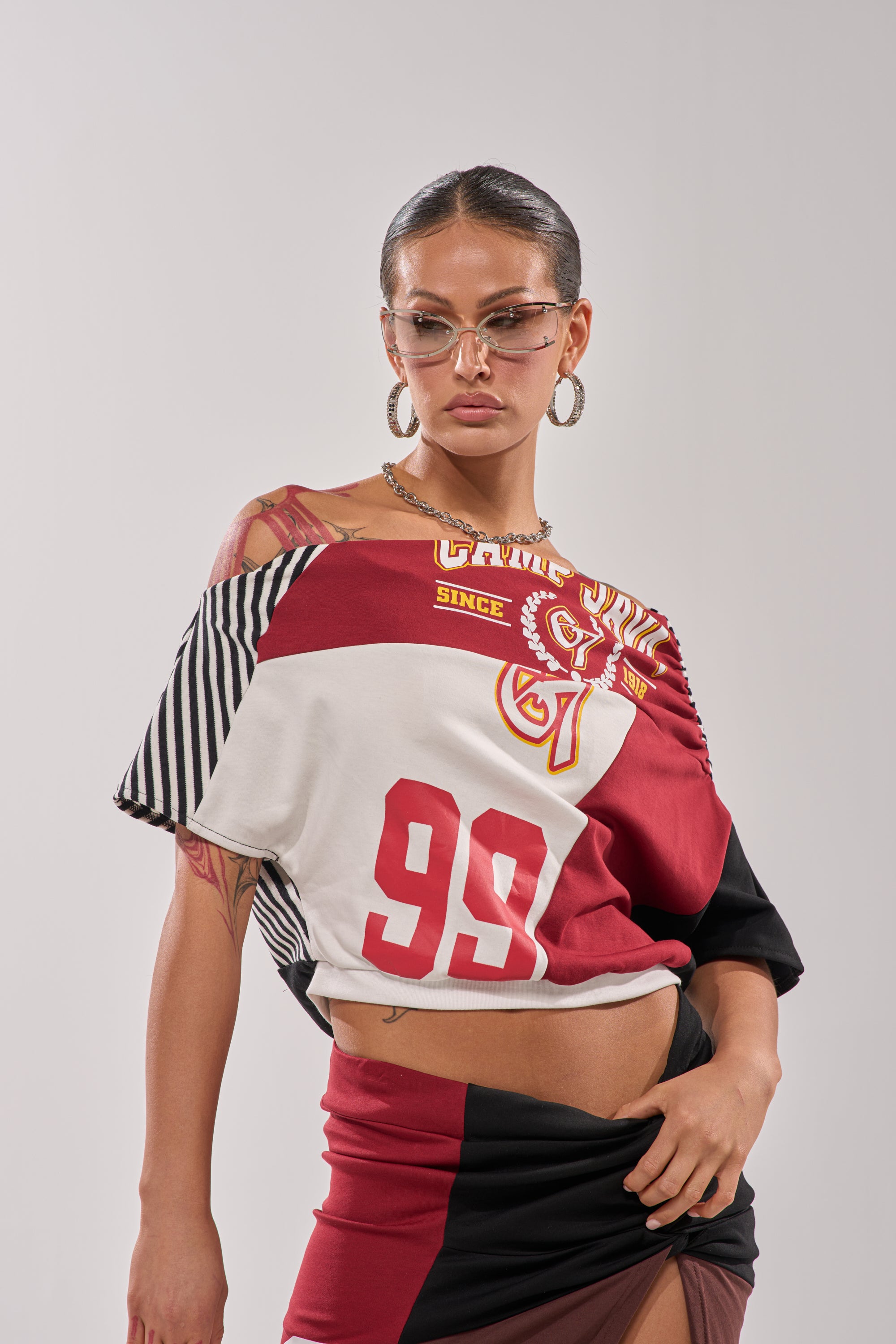A woman with glasses and hoop earrings poses confidently in the SCHOOL SPIRIT REWORKED TEE, a color-blocked, cropped shirt with varsity-style detailing and the number 99. She has tattoos and stands against a plain background. 2