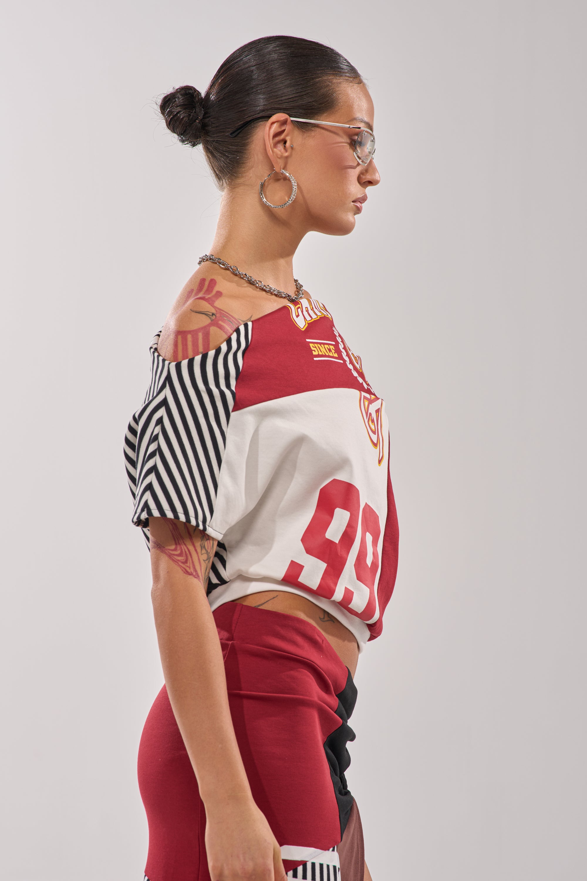 A woman with slicked-back hair, large hoop earrings, sunglasses, and visible tattoos models the SCHOOL SPIRIT REWORKED TEE—a varsity-style cropped top in red, white, and black with the number 99—exuding a streetwear vibe. 4