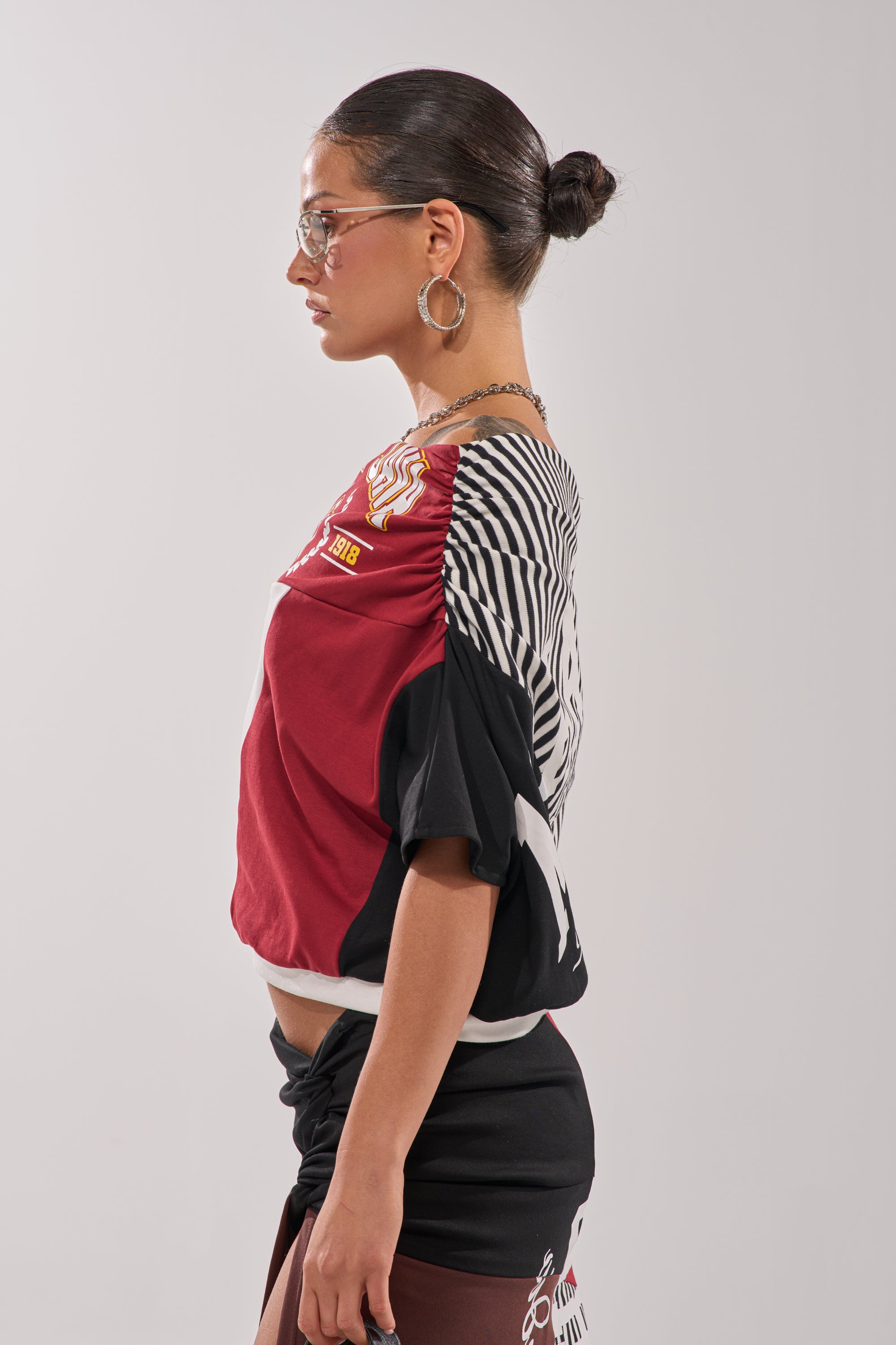 A woman with dark hair in a bun, glasses, hoop earrings, and the SCHOOL SPIRIT REWORKED TEE stands in profile against a plain light gray background. 5