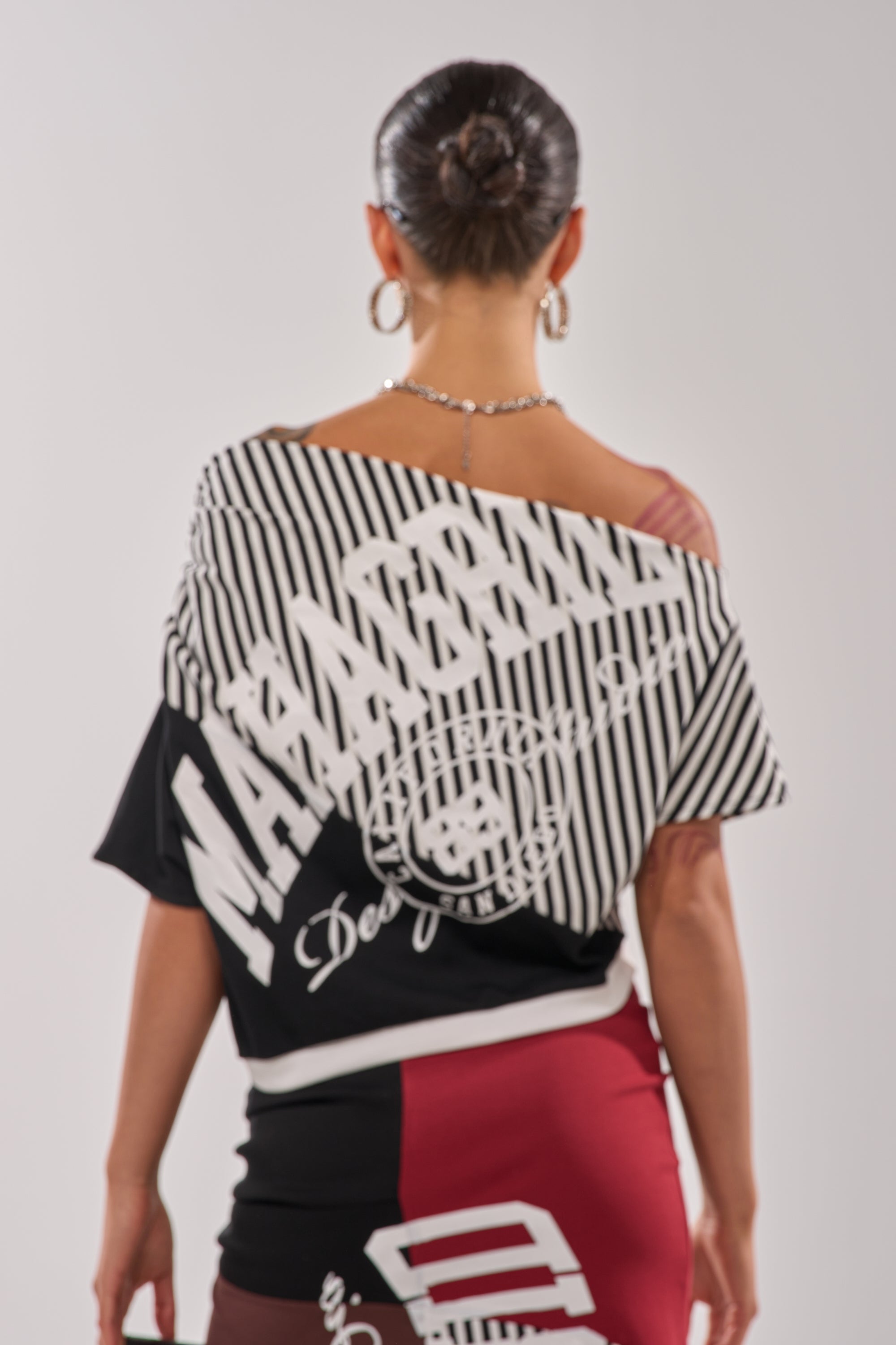 Wearing her hair in a bun and large hoop earrings, she styles the SCHOOL SPIRIT REWORKED TEE off the shoulder with a color-blocked skirt, capturing game-day flair in a back-facing pose against a simple background. 6