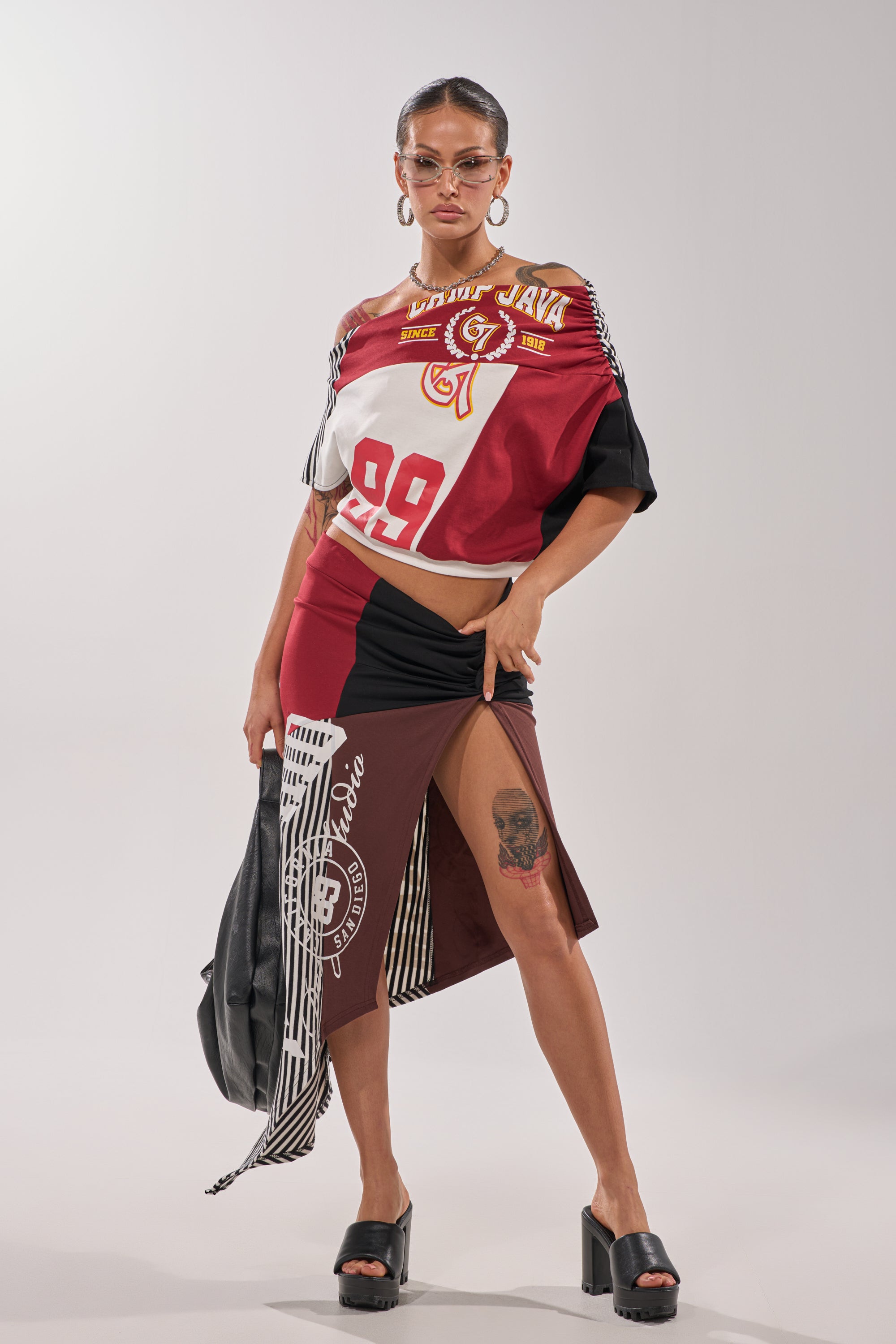 A woman poses confidently in the SCHOOL SPIRIT REWORKED TEE, styled with bold red, black, and white varsity-inspired patterns. She accessorizes with sunglasses, black heeled sandals, a jacket in hand, and displays a skull tattoo on her thigh. 7