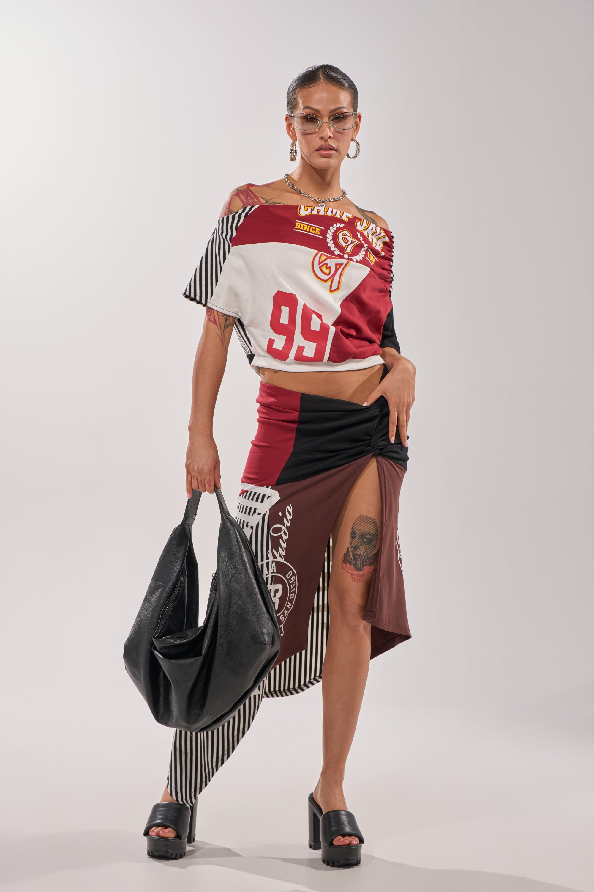 A woman poses confidently in a SCHOOL SPIRIT REWORKED TEE and a mixed-pattern, asymmetrical skirt. She accessorizes with sunglasses, hoop earrings, black platform sandals, a large black bag, and shows off her above-the-knee tattoo. 8