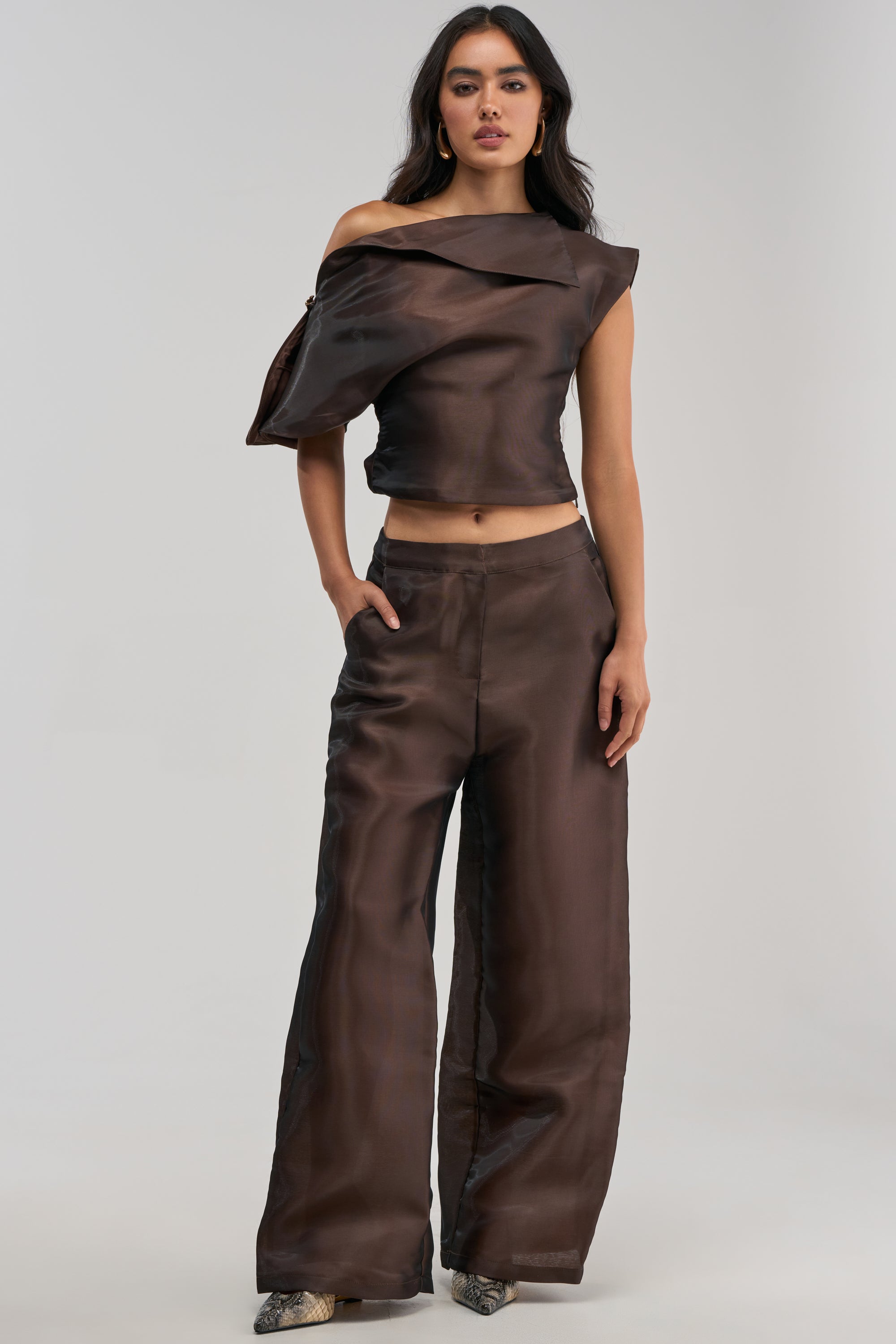 A woman stands against a plain light background, wearing the ON FIRE TROUSER IN BROWN—glossy, wide-leg trousers paired with a dark brown off-shoulder asymmetric top. With one hand in her pocket, she creates an effortlessly chic look, perfect for wedding guest styling.