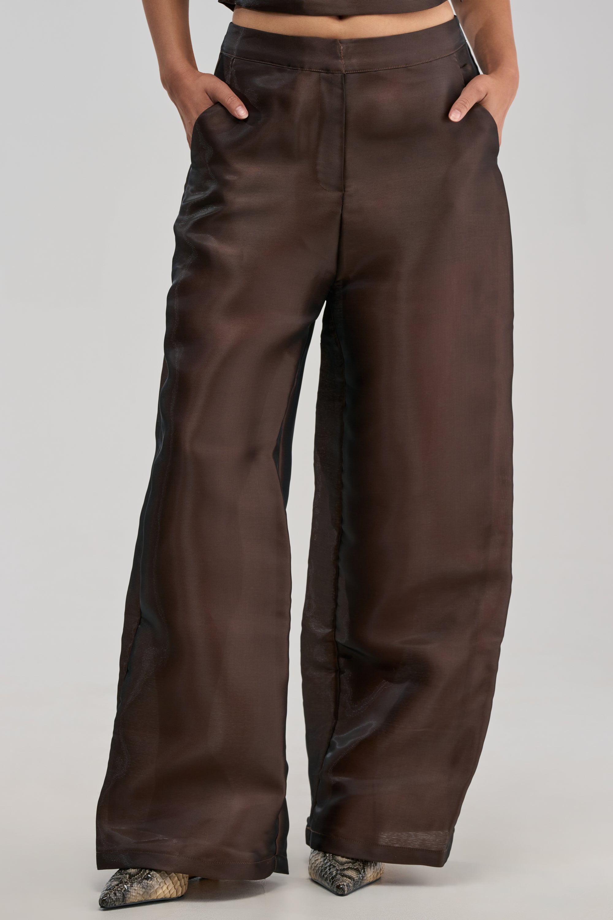 Wearing the ON FIRE TROUSER IN BROWN—a sheer, glossy wide-leg style with pockets—paired with snake-patterned shoes, this effortlessly chic look is perfect for modern wedding guest styling against a minimalist backdrop.