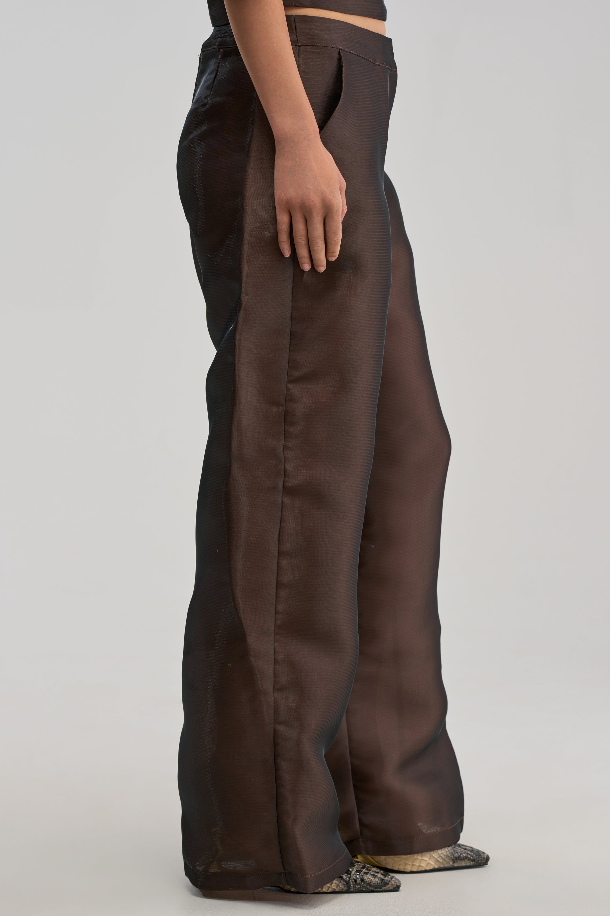 A person models the ON FIRE TROUSER IN BROWN—high-waisted, wide-leg, and glossy—paired with pointed shoes against a plain light gray background. Only their lower torso and legs are visible.