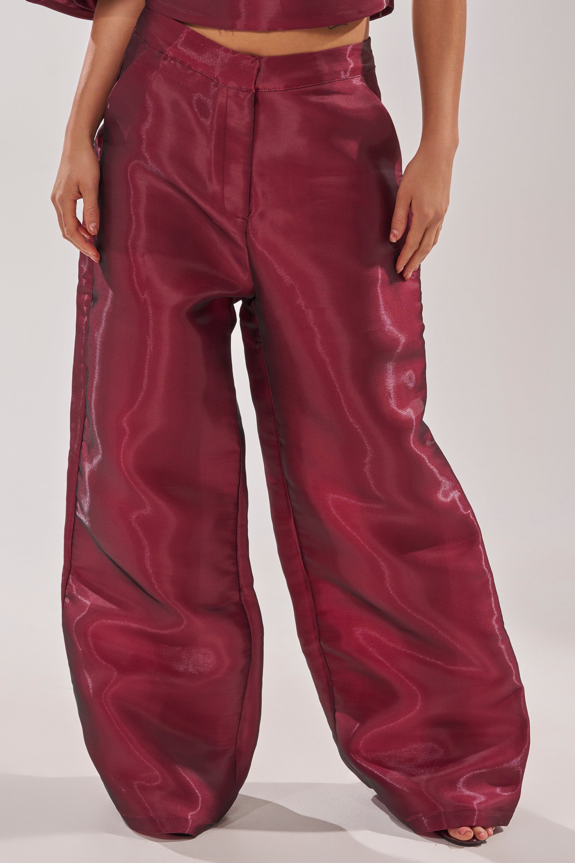 A person stands barefoot against a plain background, wearing the ON FIRE TROUSER—high-rise wide-leg pants in shiny burgundy satin—making these statement trousers the highlight of their effortlessly stylish look. 1