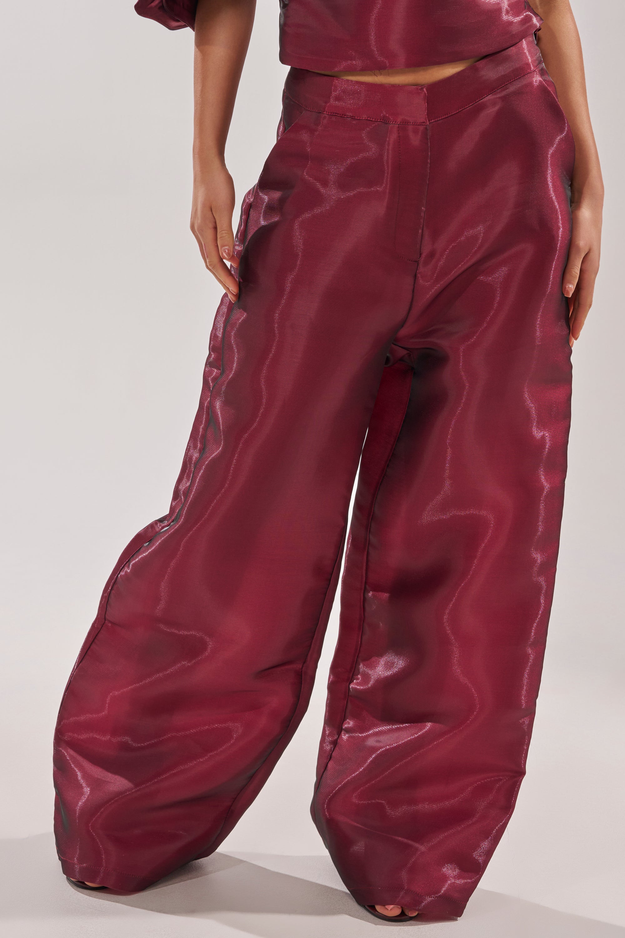 A person wears the ON FIRE TROUSER—shiny, high-rise, wide-leg burgundy pants with a loose fit. Only the lower half is shown against a plain background, drawing attention to these bold statement trousers. 2