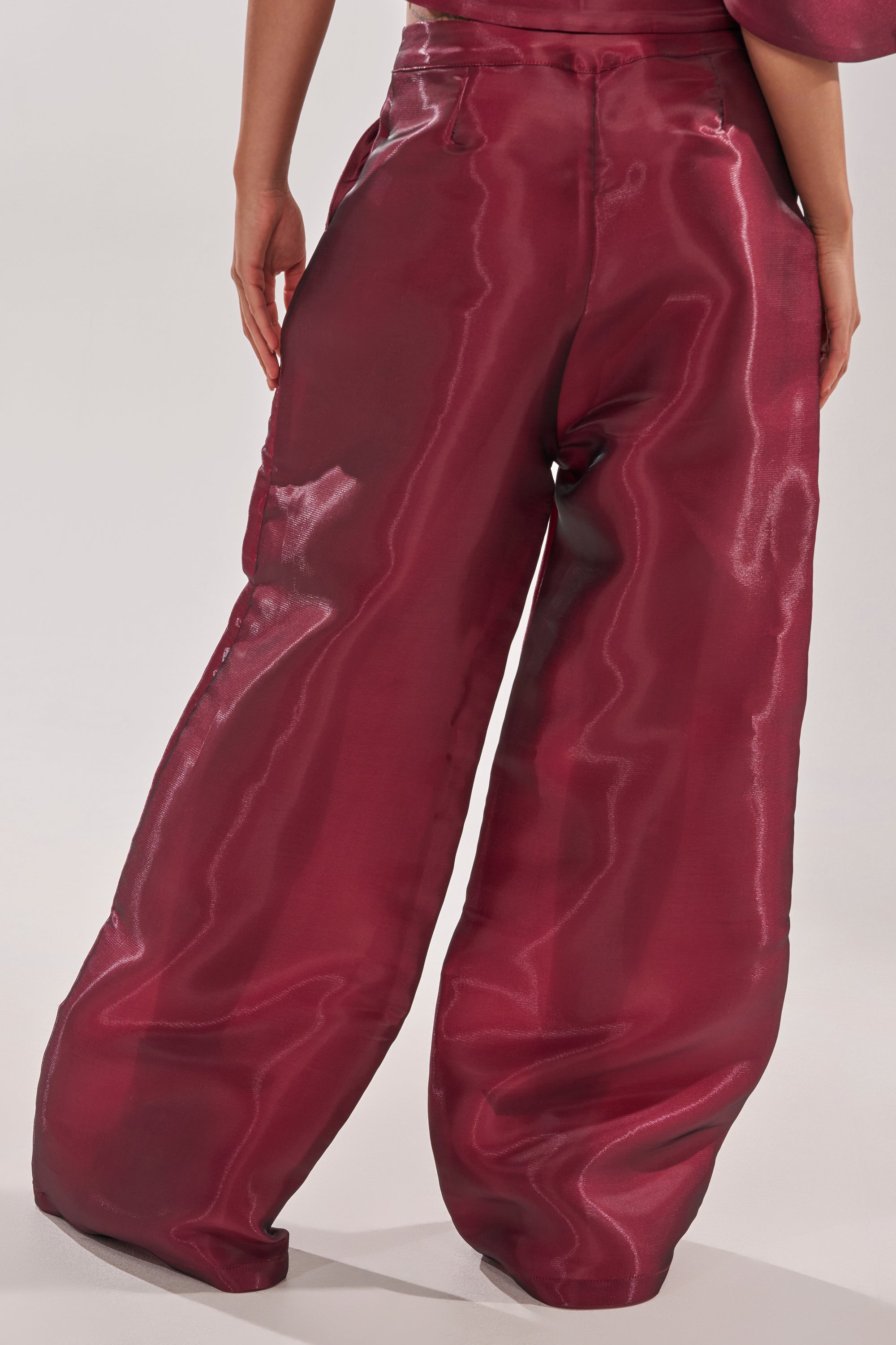 A person wearing the ON FIRE TROUSER, a high-rise wide-leg burgundy pant with a glossy finish, stands with their back to the camera against a plain light background. 4