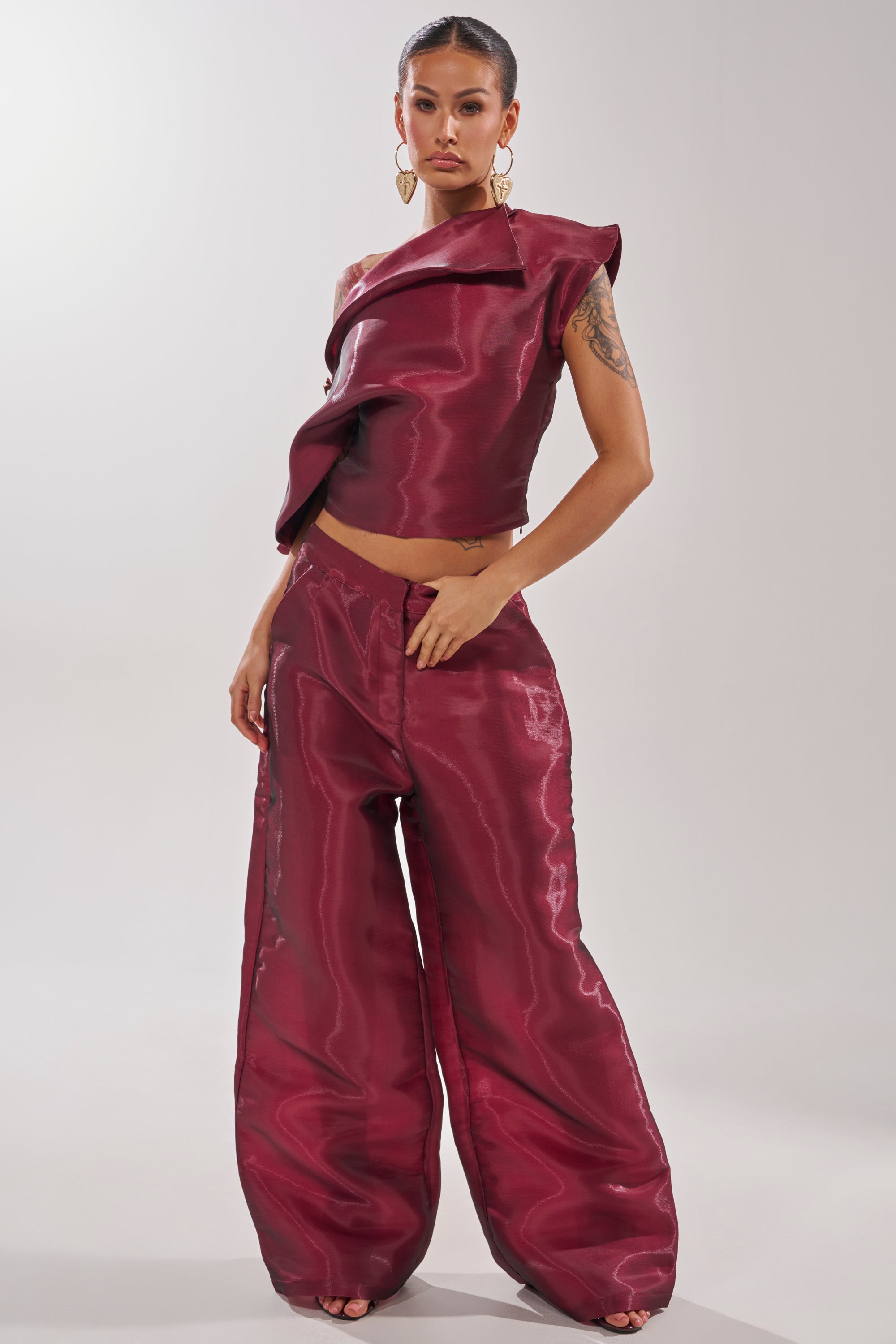 A woman models the ON FIRE TROUSER, a shiny burgundy high-rise wide-leg pant paired with a one-shoulder asymmetrical top. She accessorizes with large gold earrings and has her hair pulled back against a plain light background. 5