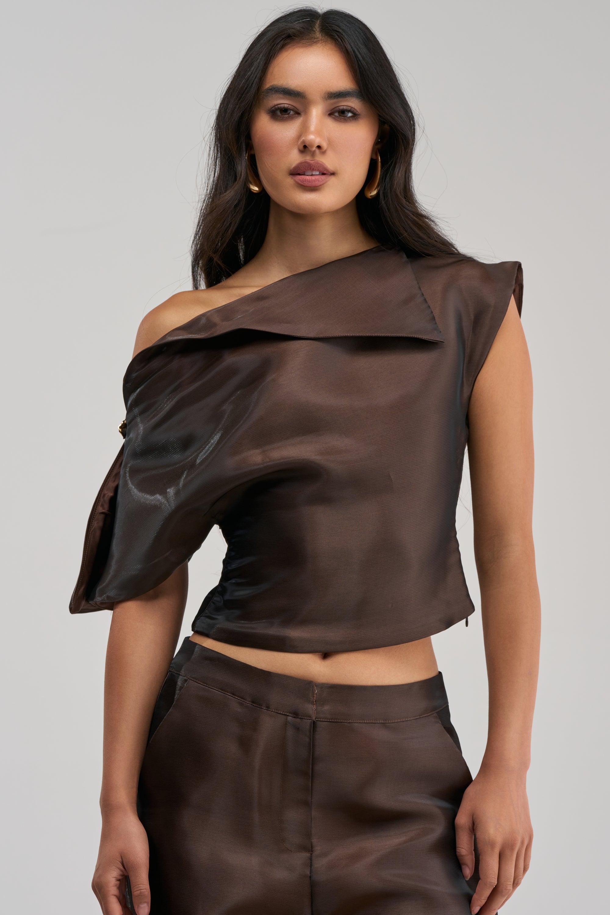 A woman wearing the ON FIRE BLOUSE IN BROWN, featuring a glossy finish and an asymmetrical off-the-shoulder sleeve, with matching pants, stands against a plain background.