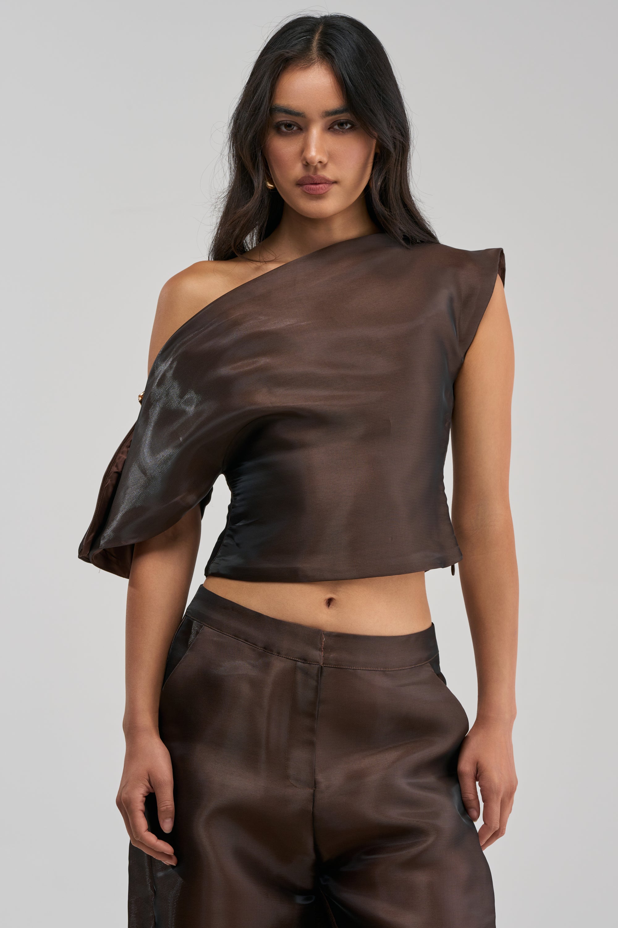 A woman stands facing forward, wearing the ON FIRE BLOUSE IN BROWN—a glossy, asymmetrical off-the-shoulder top—paired with matching high-waisted pants. She has long, wavy dark hair and a neutral expression, set against a plain light gray background.