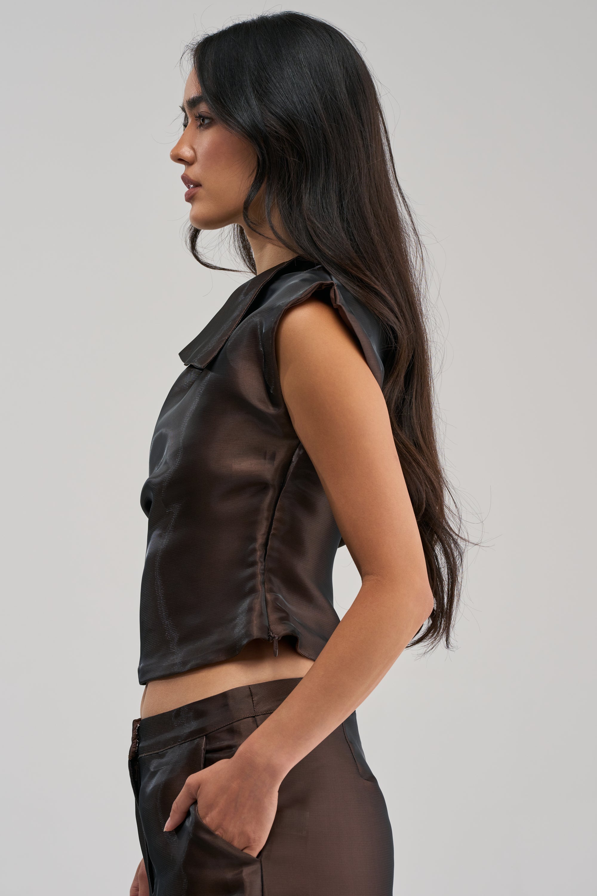 A woman with long dark hair stands in profile, wearing the ON FIRE BLOUSE IN BROWN and matching brown pants against a plain light gray background.