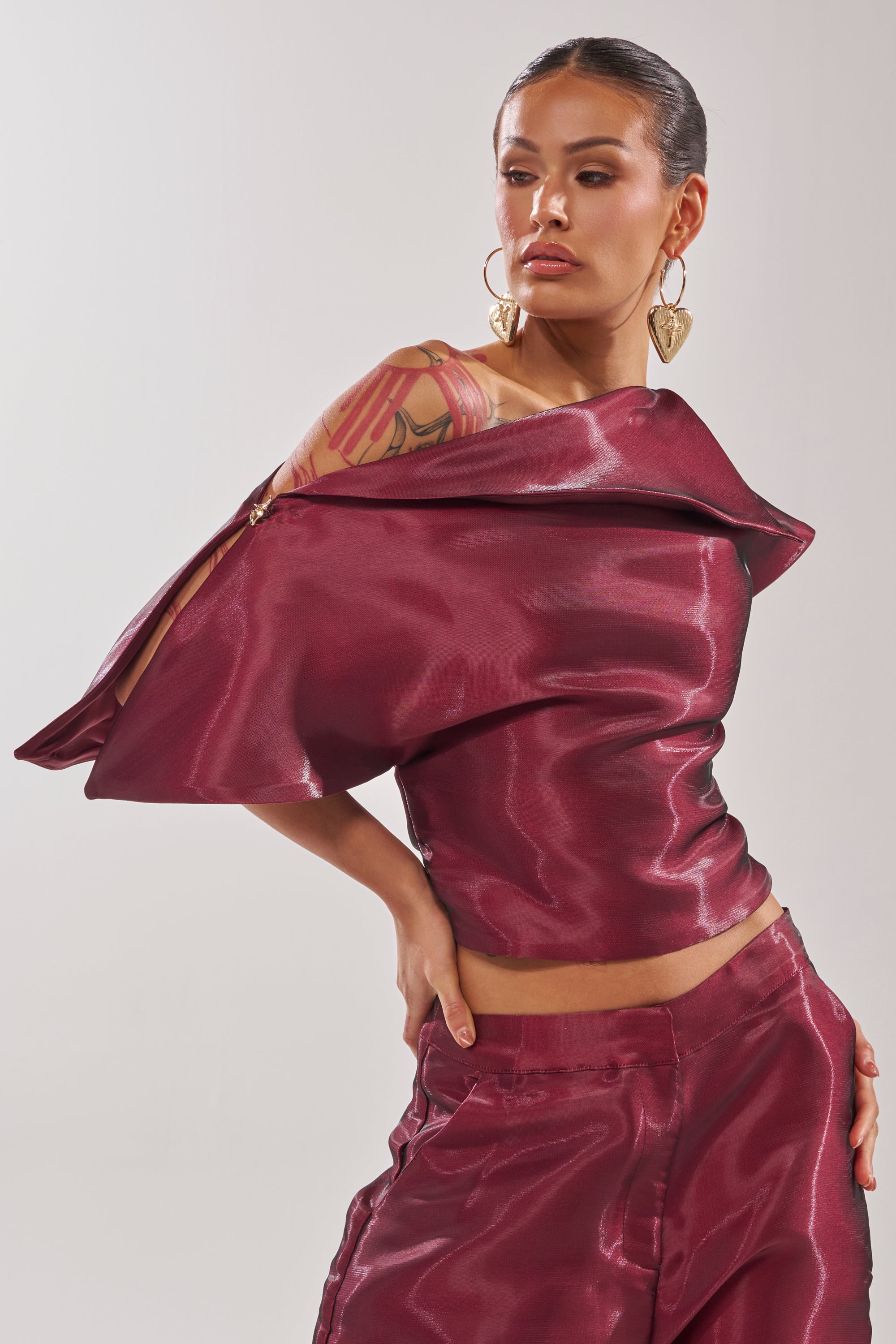 A person models the ON FIRE BLOUSE—a glossy burgundy, off-the-shoulder top with dramatic, angular sleeves—paired with matching pants. They wear large hoop earrings, reveal tattoos, and have slicked-back hair. 0