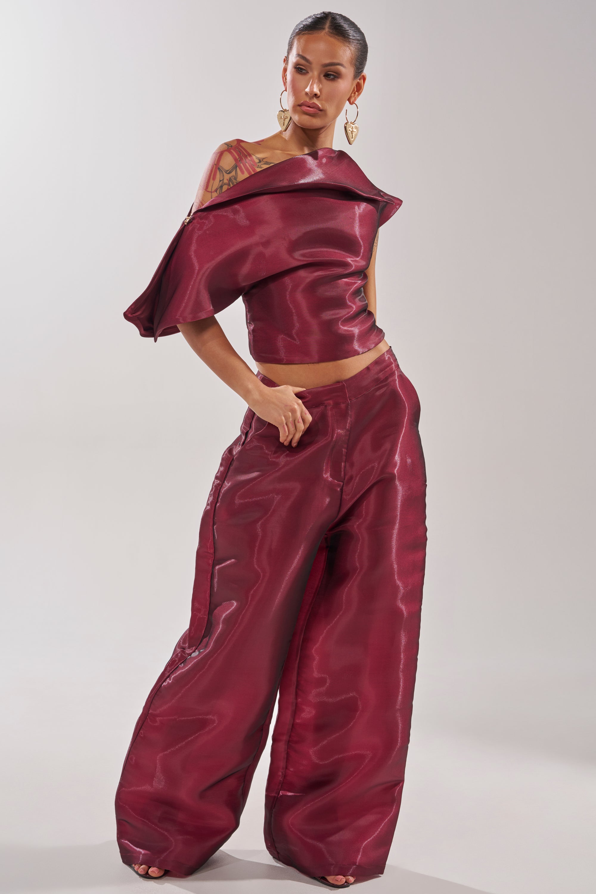 A woman models the ON FIRE BLOUSE, a shiny dark red off-the-shoulder top with an asymmetrical neckline, paired with matching wide-leg pants and bold gold earrings. Her hair is pulled back as she stands against a plain light background. 9