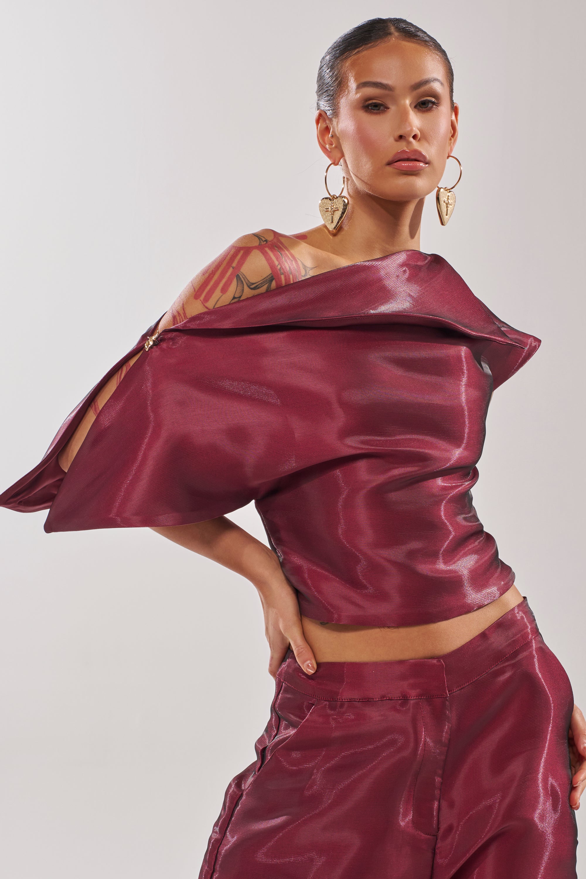 A woman models the ON FIRE BLOUSE, a burgundy asymmetrical top with a glossy finish, paired with matching pants. She poses with one hand on her hip, showcasing gold heart earrings and a floral tattoo on her upper arm. 1