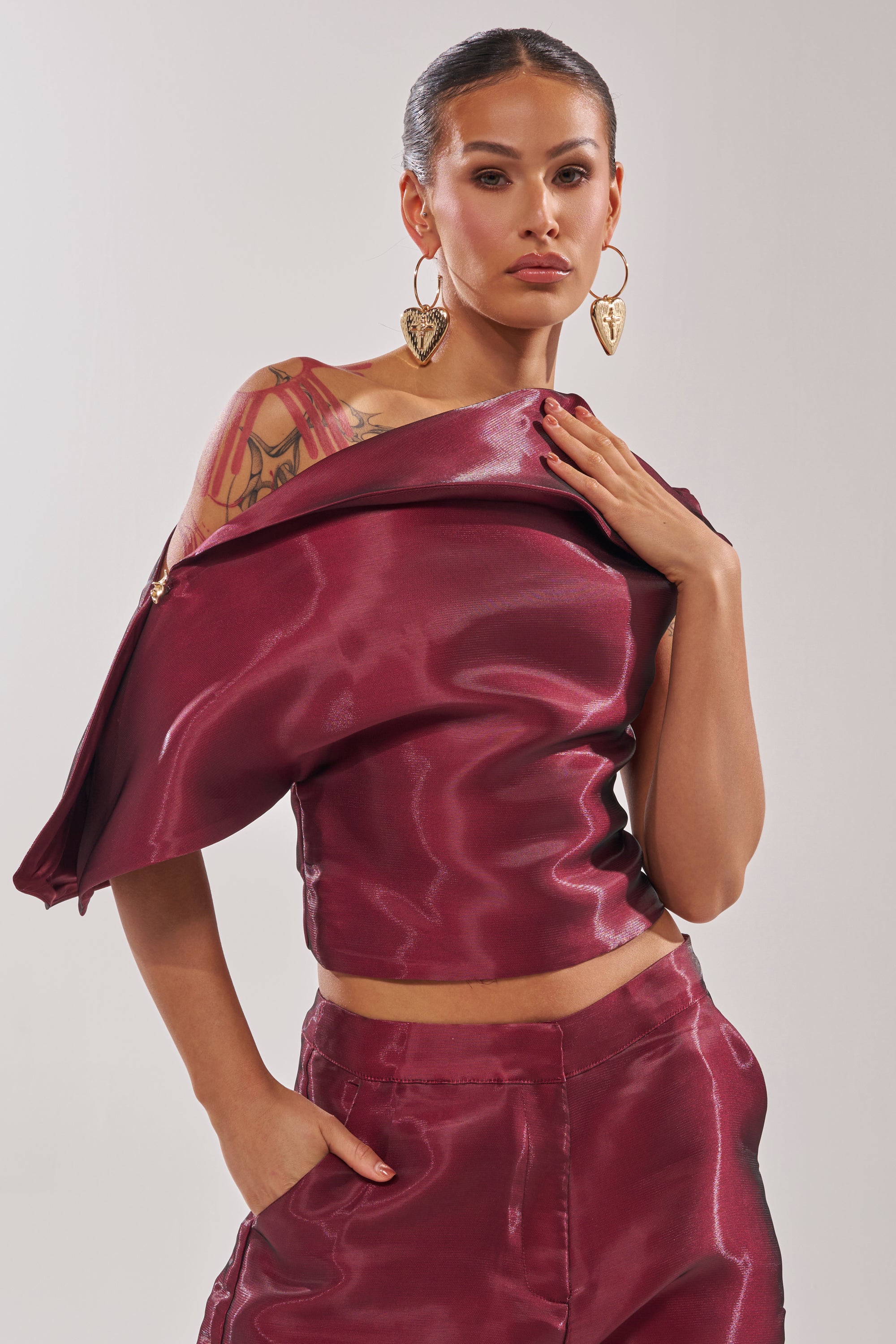 Wearing the ON FIRE BLOUSE in shiny burgundy with matching pants, a person poses with one hand in their pocket and the other on their collar, showcasing large leaf-shaped earrings and a visible arm tattoo. 2