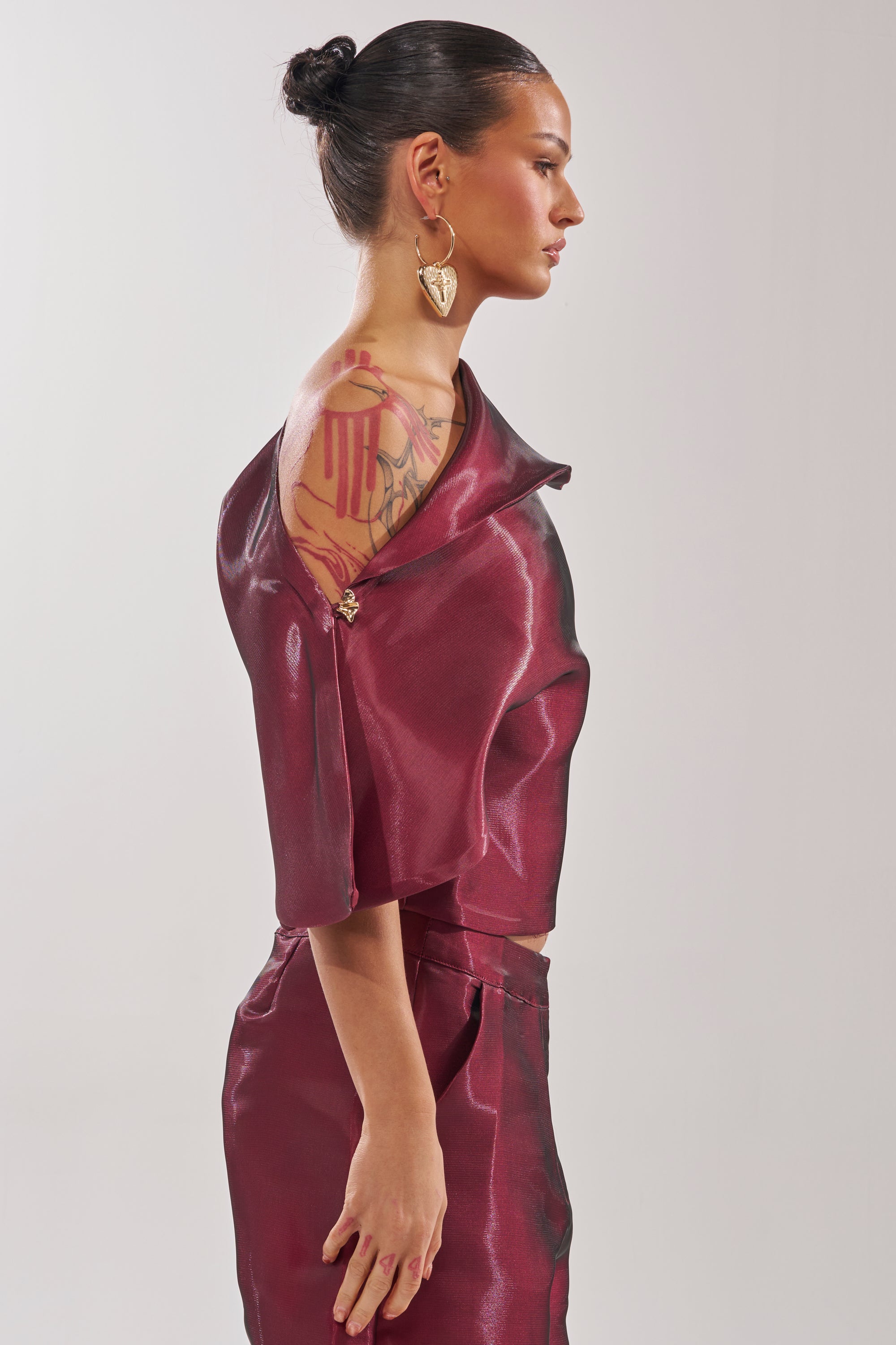 A woman with her hair in a bun models the ON FIRE BLOUSE, a shiny burgundy off-the-shoulder top revealing a colorful arm tattoo. She faces left in a neutral pose against a plain light background, wearing a large heart-shaped earring. 4
