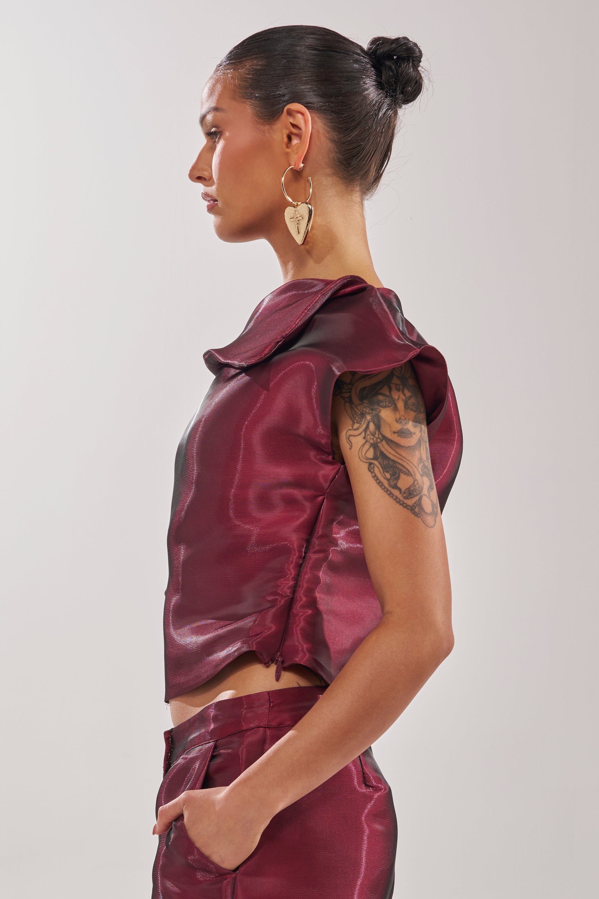 A woman with dark hair in a bun, gold heart earrings, and a detailed upper arm tattoo is shown in profile wearing the sleeveless, shiny maroon ON FIRE BLOUSE. 5