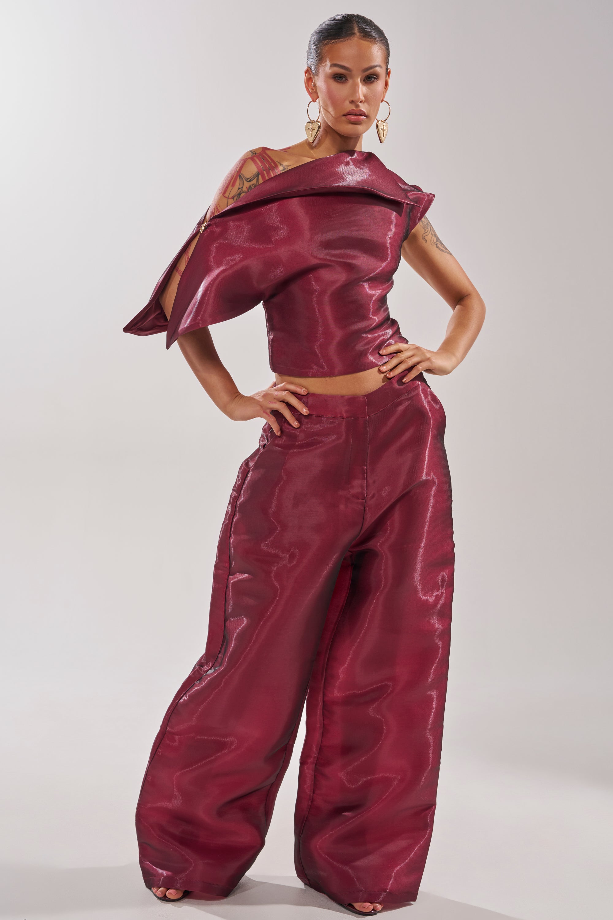 A woman poses confidently in the ON FIRE BLOUSE, a shiny dark red top with sculptural shoulder detail, paired with matching wide-leg pants. She accessorizes with large hoop earrings and has slicked-back hair and visible tattoos. 7