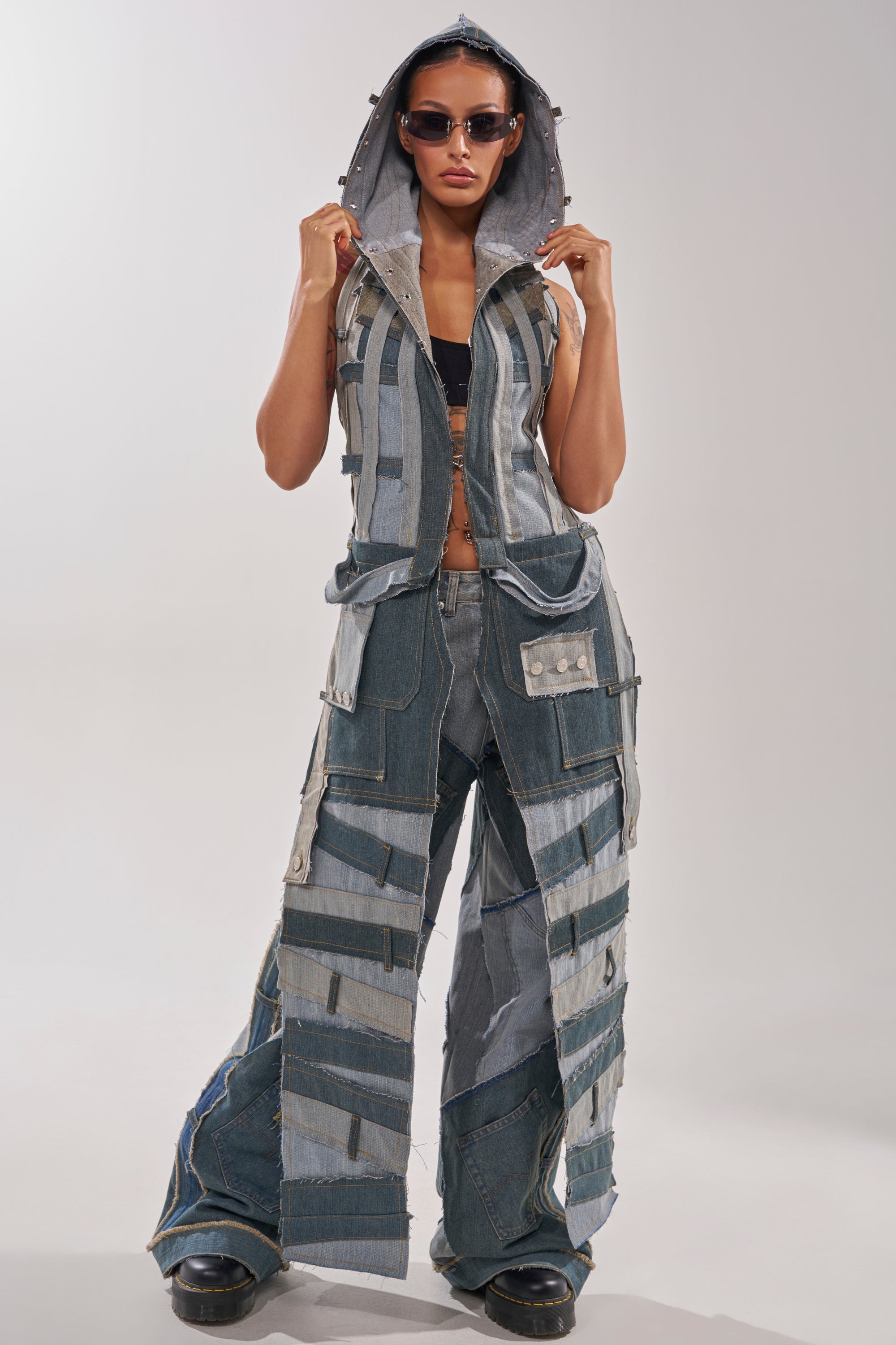 A woman exudes street style in sunglasses and black boots, wearing the ALL THAT DISTRESSED HOODED TOP—a patchwork denim sleeveless hoodie with frayed edges, visible stitching, and various blue shades—matched with wide-leg pants. 0