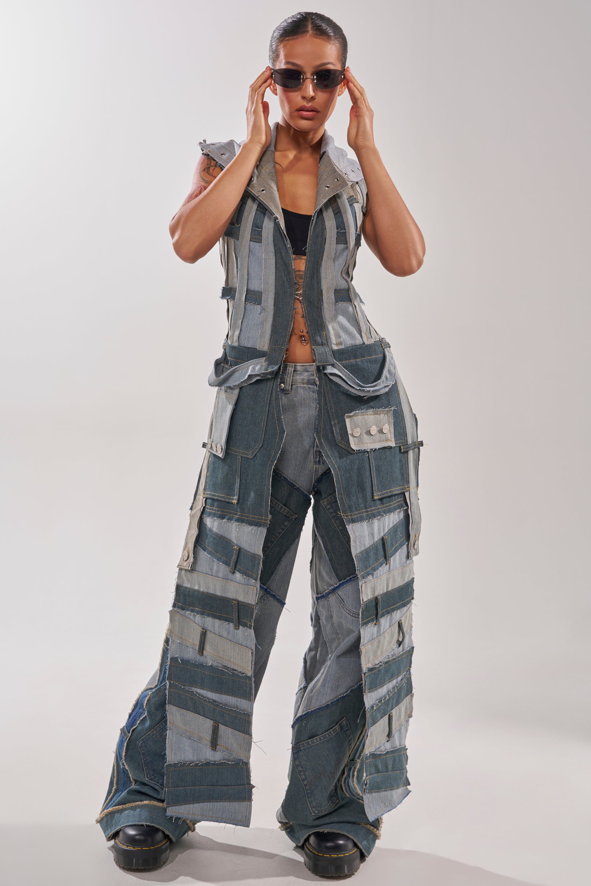 Wearing the ALL THAT DISTRESSED HOODED TOP, a woman channels street-style energy as she adjusts her sunglasses, pairing the edgy hoodie with statement jeans and a black bra top against a plain background. 1