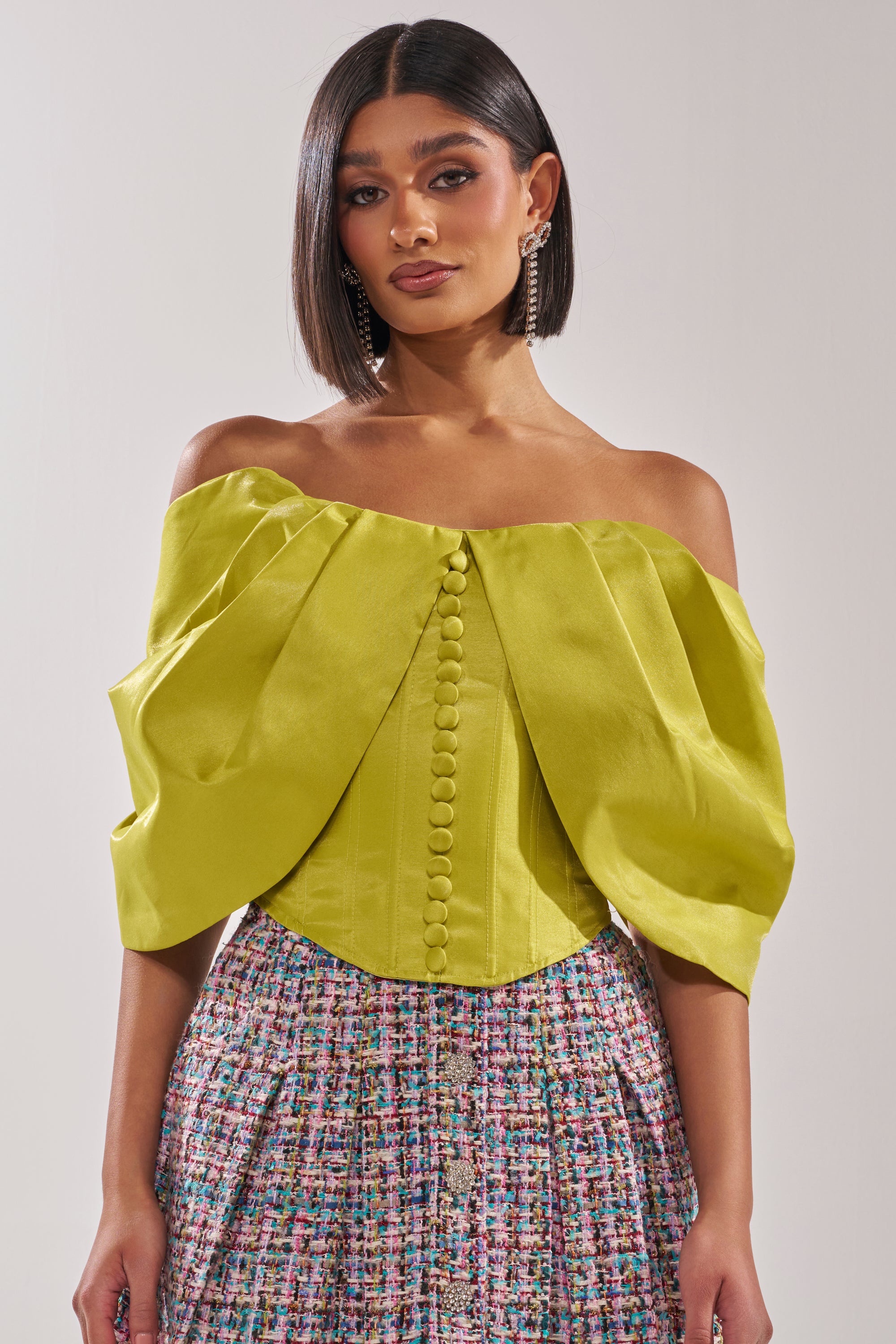 A woman with straight, shoulder-length hair wears the ELECTRIC CORSET IN GREEN styled off-the-shoulder with puff sleeves and front buttons, paired with a high-waisted, multicolored patterned skirt. 0