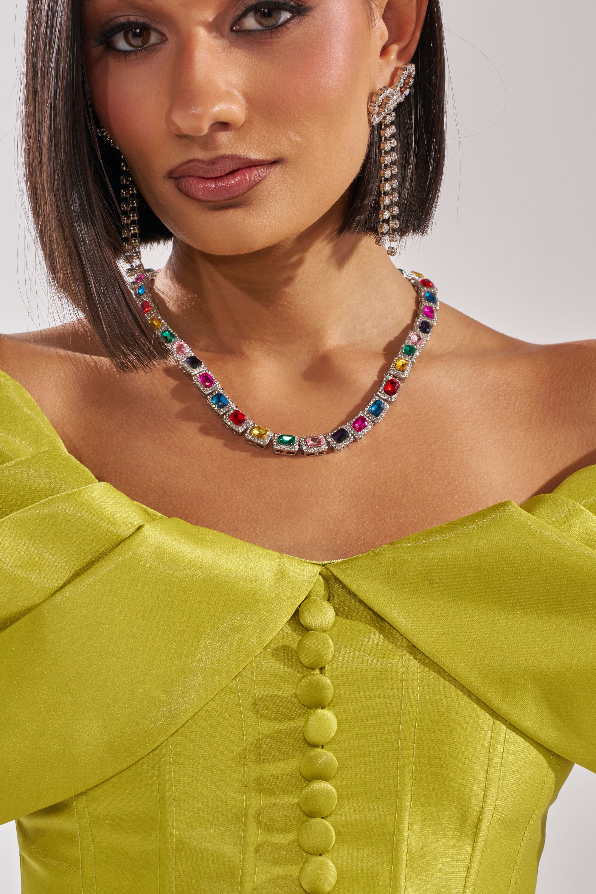 A woman in the ELECTRIC CORSET IN GREEN, an off-shoulder dress with fabric-covered buttons, pairs it with a colorful gemstone necklace and sparkly chain ear cuff. Her sleek bob and straight dark hair complete the look. 9