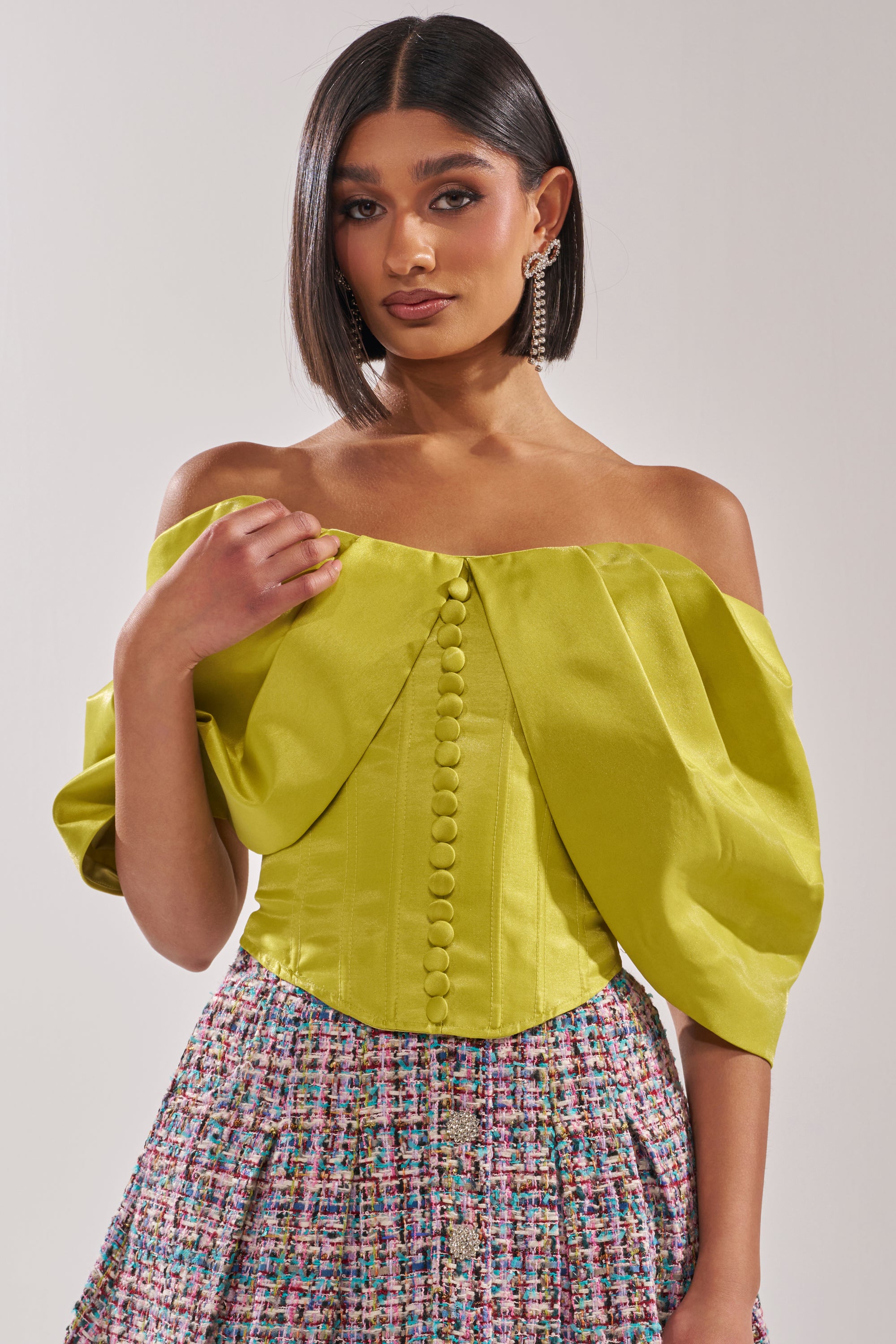 A woman with straight, dark hair wears the ELECTRIC CORSET IN GREEN, styled with a textured, multicolored skirt for a couture-inspired silhouette against a plain light background. 1