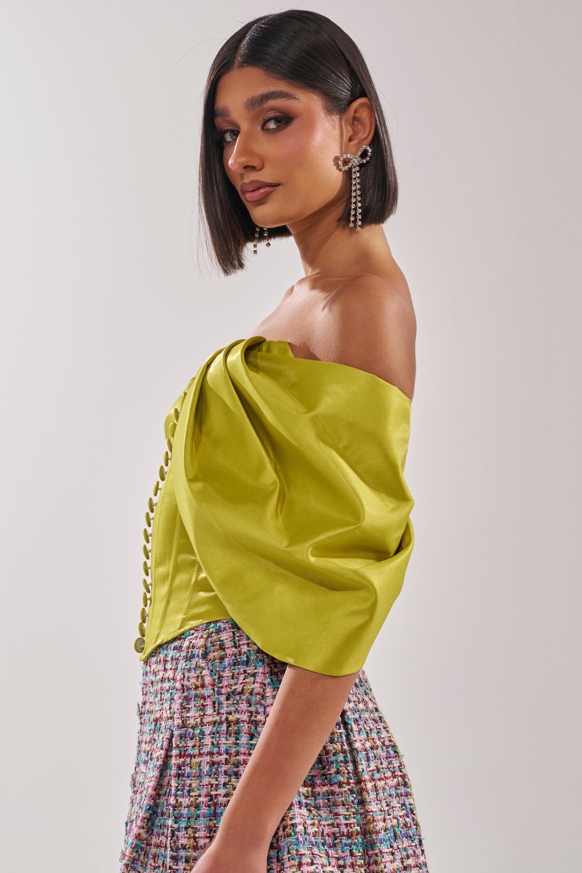 A woman with straight, shoulder-length dark hair wears the ELECTRIC CORSET IN GREEN with decorative buttons and voluminous sleeves, paired with a colorful textured skirt. She completes the look with statement earrings and a confident expression. 3