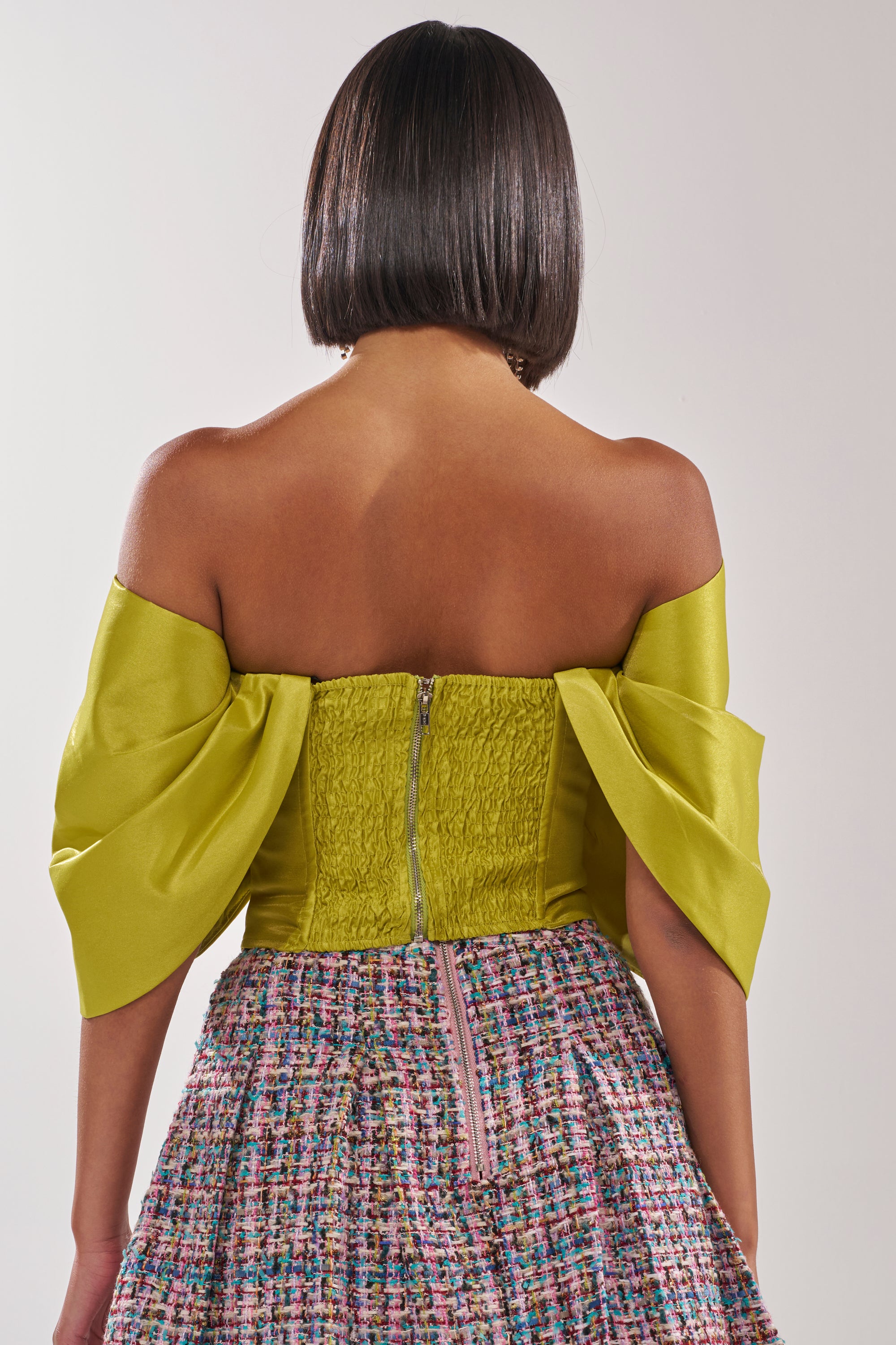 A person with straight, chin-length hair is shown from the back, wearing the ELECTRIC CORSET IN GREEN paired with a multicolored tweed skirt, both featuring visible zippers. 4