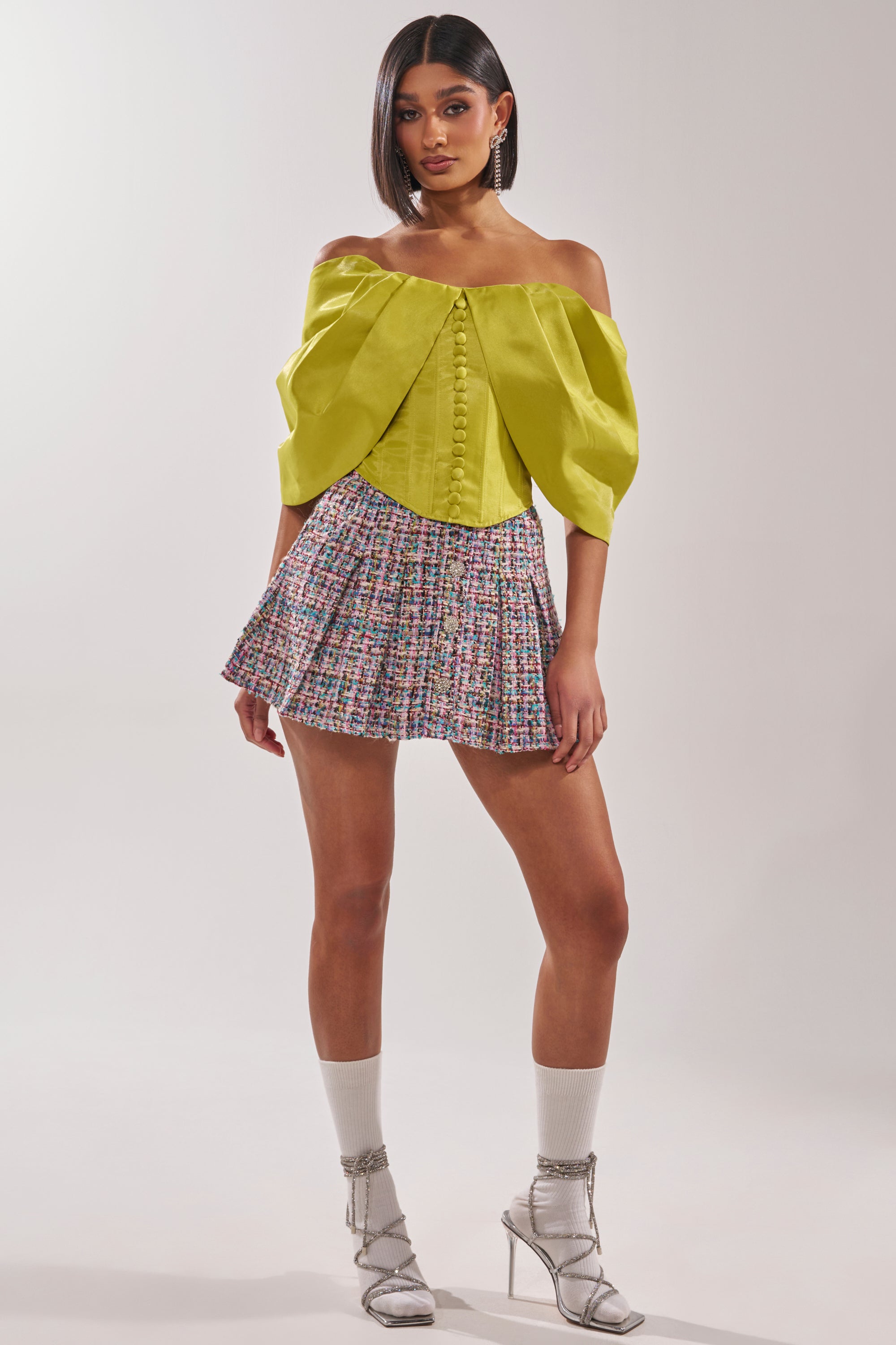 A woman stands wearing the ELECTRIC CORSET IN GREEN with puff sleeves, a multicolored pleated mini skirt, silver high-heeled sandals, and light-colored socks against a plain background. 5