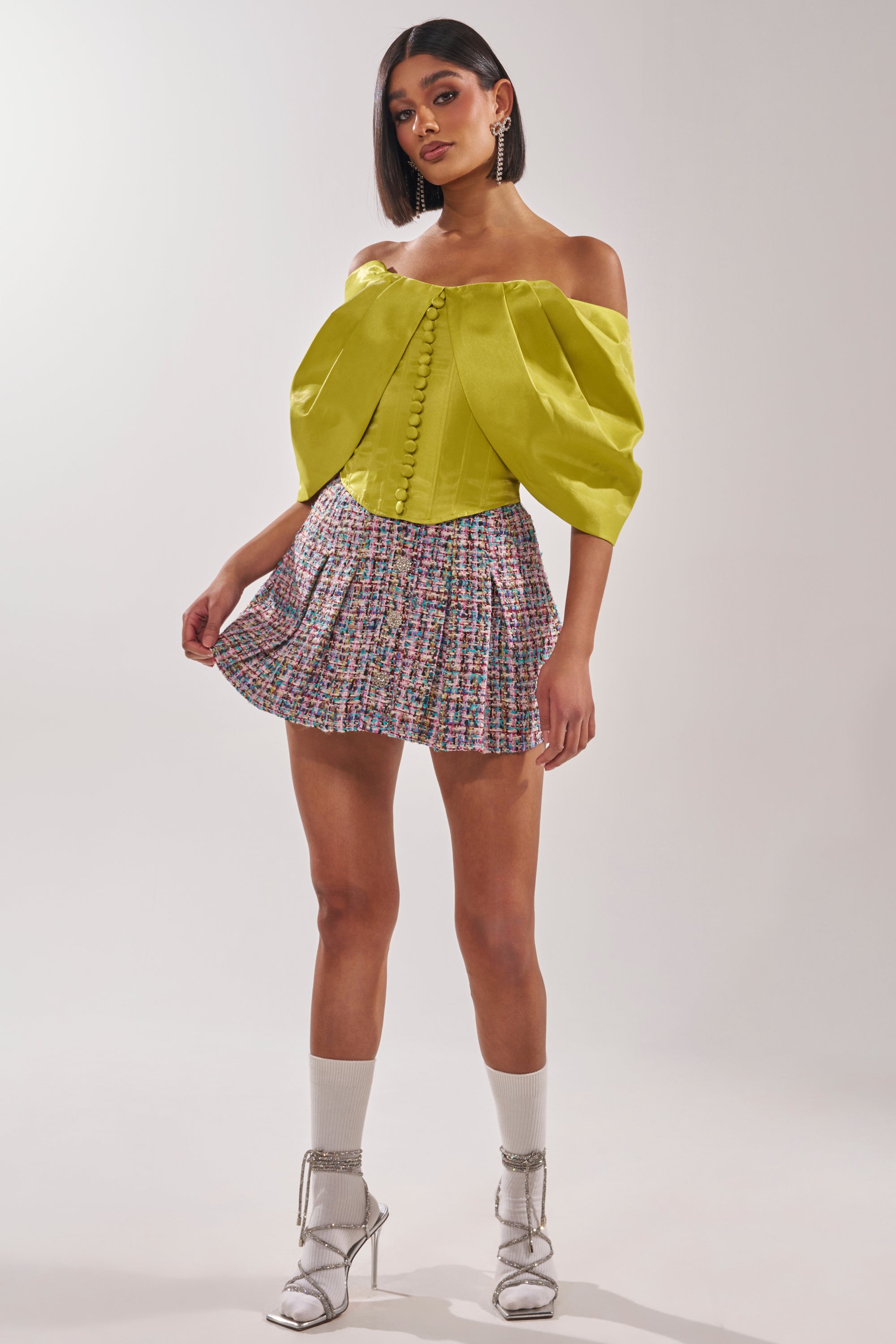 A woman poses in the ELECTRIC CORSET IN GREEN with front buttons, styled with a colorful pleated mini skirt, silver heels, white socks, and drop earrings against a plain light background. 6