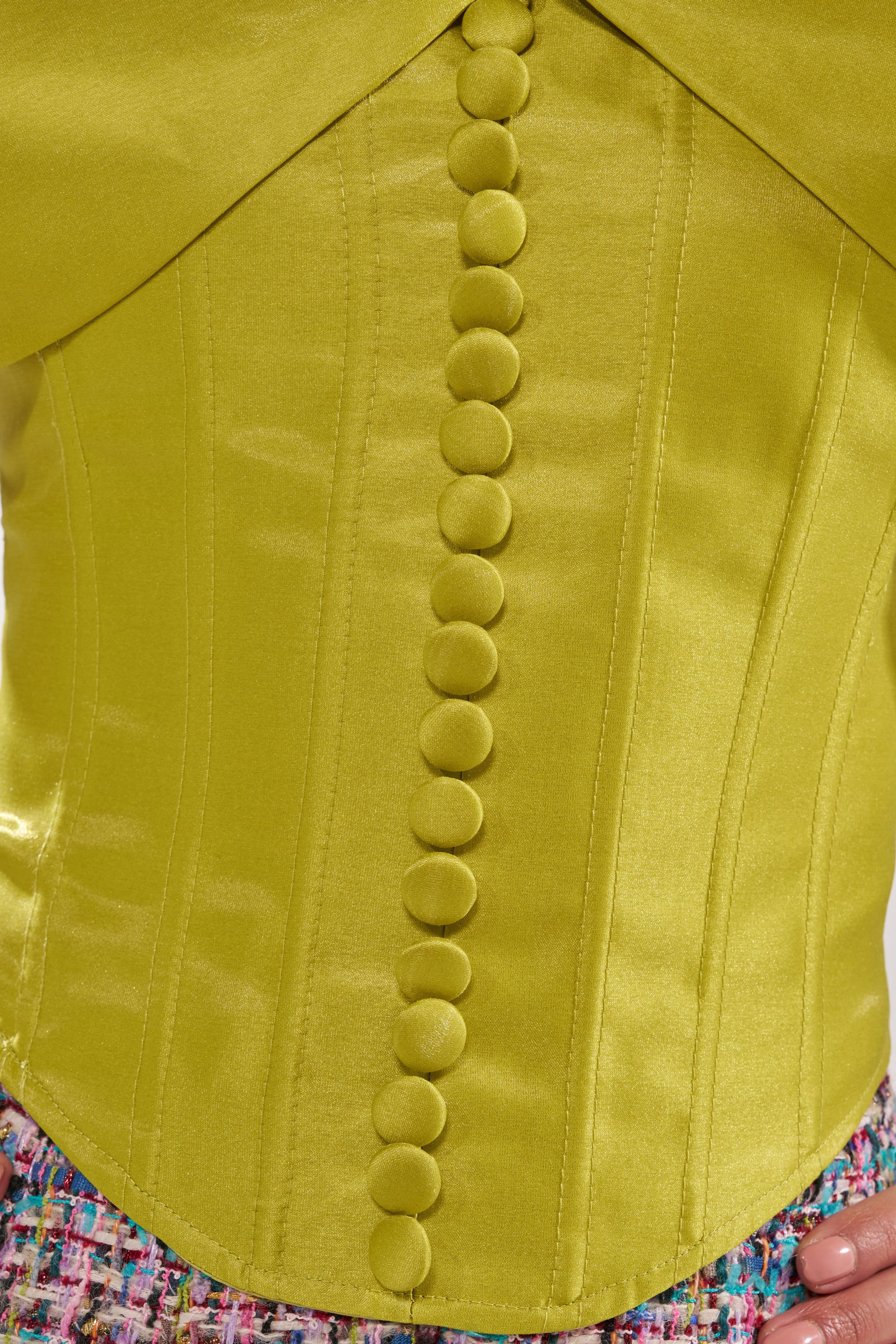 Close-up of the ELECTRIC CORSET IN GREEN featuring fabric-covered buttons in a single row, styled over a multicolored textured skirt. A manicured hand rests at the edge of the frame. 8