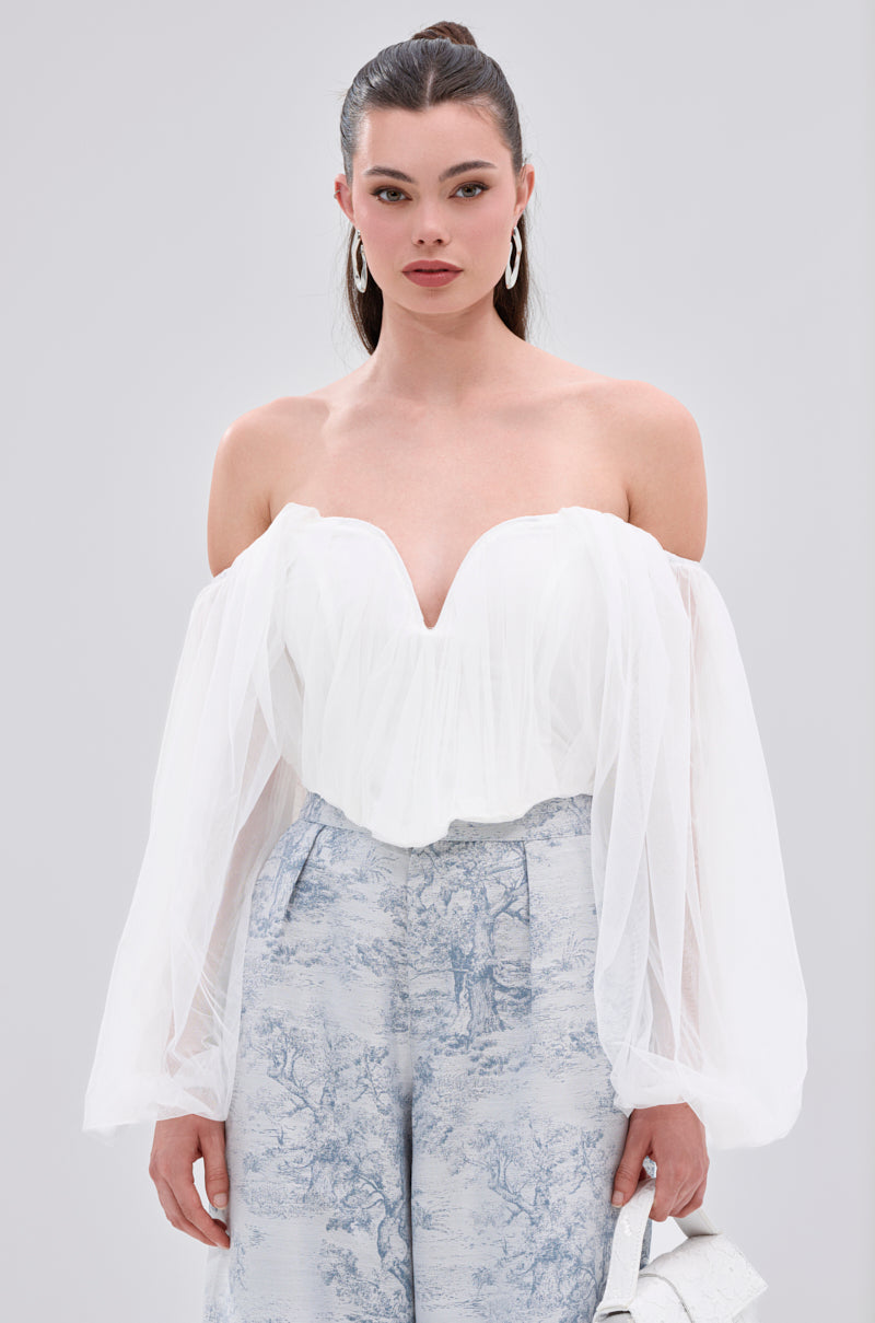 A woman wears the FLY WITH ME CORSET TOP IN WHITE, an off-the-shoulder piece with sheer long sleeves and a deep sweetheart neckline, styled with light blue high-waisted pants and a light-colored handbag.