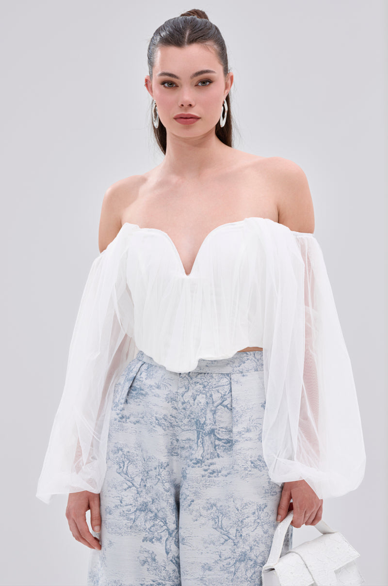 A woman wears the FLY WITH ME CORSET TOP IN WHITE with organza sleeves, paired with high-waisted blue and white patterned pants. She stands against a plain background, holding a small white handbag and wearing hoop earrings.