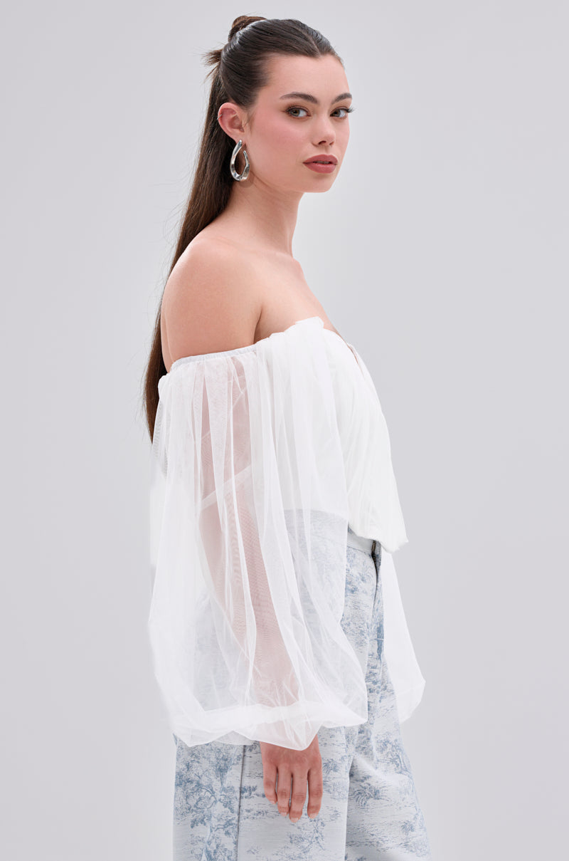 A woman with long dark hair in a half-up style wears the FLY WITH ME CORSET TOP IN WHITE with sheer organza sleeves and light blue patterned pants, standing sideways against a plain light gray background.
