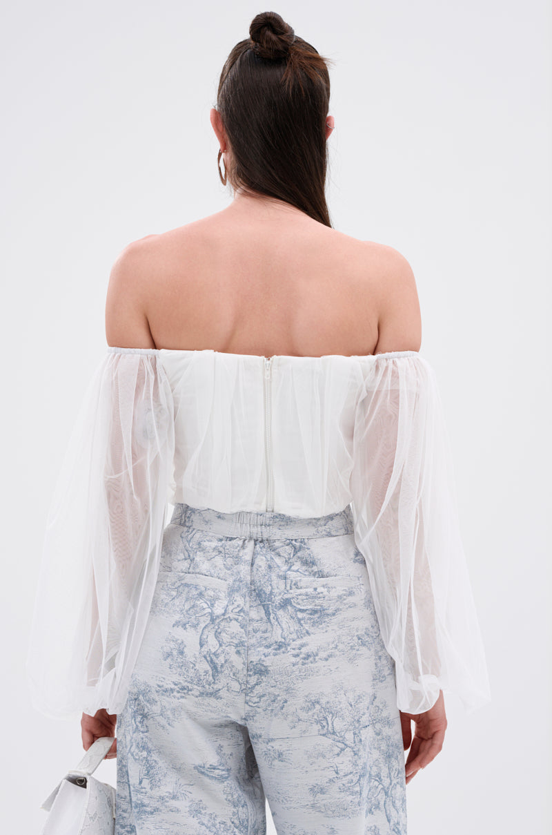 A woman with long hair half up in a bun is seen from behind wearing the FLY WITH ME CORSET TOP IN WHITE, featuring sheer organza sleeves and a sweetheart neckline, paired with high-waisted light blue toile-printed pants and holding a small white bag.