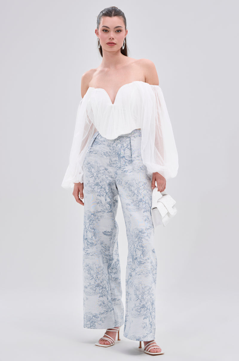 A woman wears the FLY WITH ME CORSET TOP IN WHITE, featuring a sweetheart neckline and billowy organza sleeves, styled with high-waisted, wide-leg light blue patterned pants, a small white clutch, and heeled sandals against a plain background.