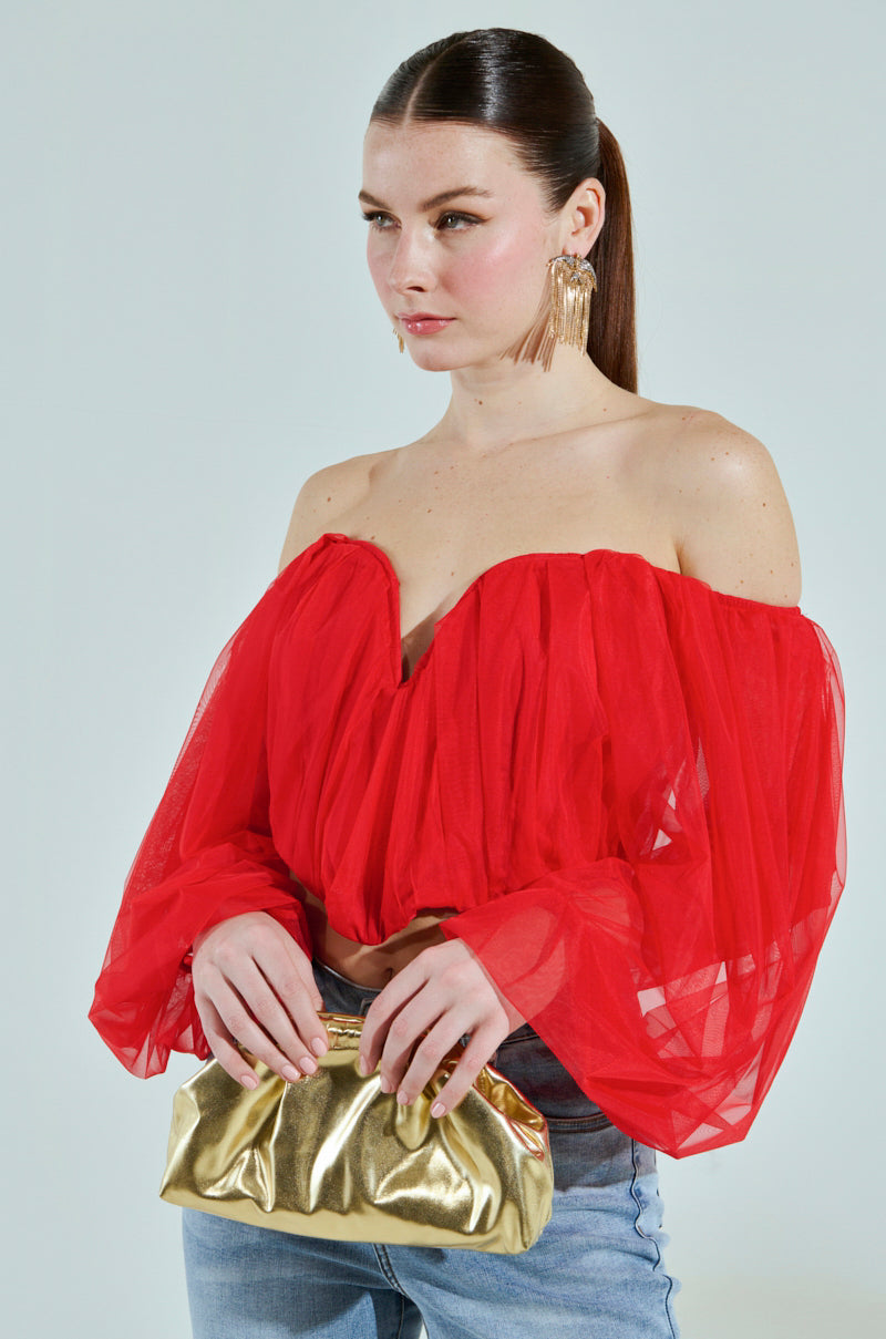 A person with long brown hair tied back wears an off-the-shoulder FLY WITH ME CORSET TOP and large dangling earrings. They hold a shiny gold clutch purse and are dressed in light blue jeans, set against a plain light background.