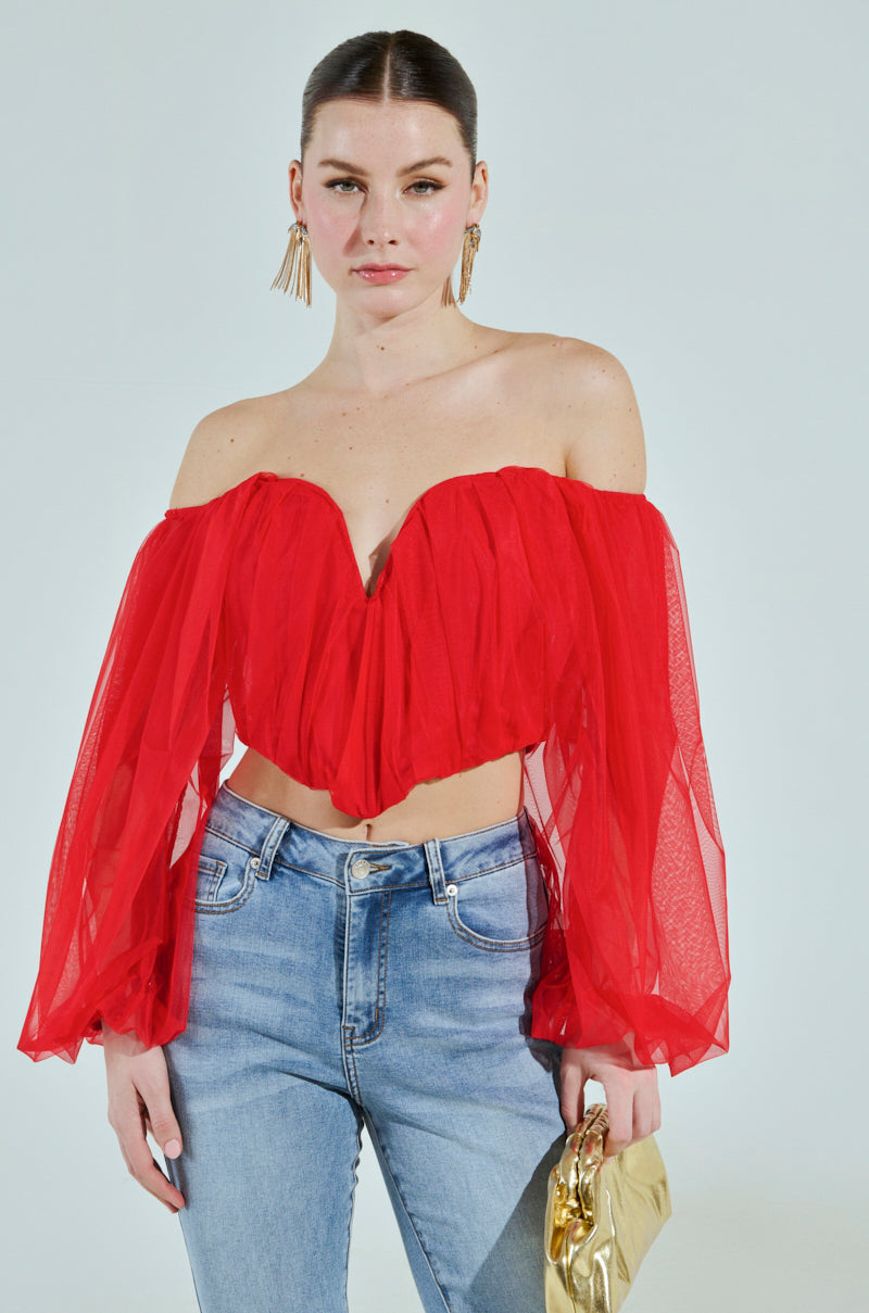 A woman wearing the FLY WITH ME CORSET TOP, a red off-the-shoulder piece, pairs it with blue jeans and a gold clutch. Her long earrings catch the light, while her hair is styled back elegantly as she poses against a plain light background.
