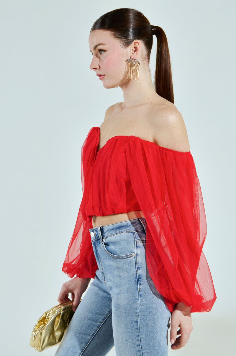 A person with long hair in a ponytail is wearing the FLY WITH ME CORSET TOP, styled as a red off-the-shoulder sheer long-sleeve top, paired with light blue jeans. They hold a gold clutch and wear large gold earrings, posing against a plain backdrop.