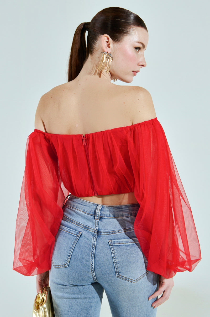A person with long brown hair in a ponytail wears the FLY WITH ME CORSET TOP, a sheer red off-the-shoulder design with puffy sleeves, paired with blue jeans. They hold a gold clutch and wear gold earrings against a plain light gray background.